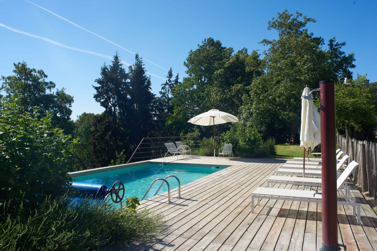An outdoor pool area photographed in wide shot during daytime, showing no people or wedding activity. The rectangular pool has turquoise water, metal entry ladders, and a partially rolled pool cover with a manual winding mechanism on one end. The pool is bordered by a weathered wooden deck furnished with white sun loungers, white metal garden chairs, an open cream parasol, and a closed cream parasol mounted on a dark red post. A rustic wooden fence runs along the right side, and the surrounding garden is densely planted. Potential venue feature image.