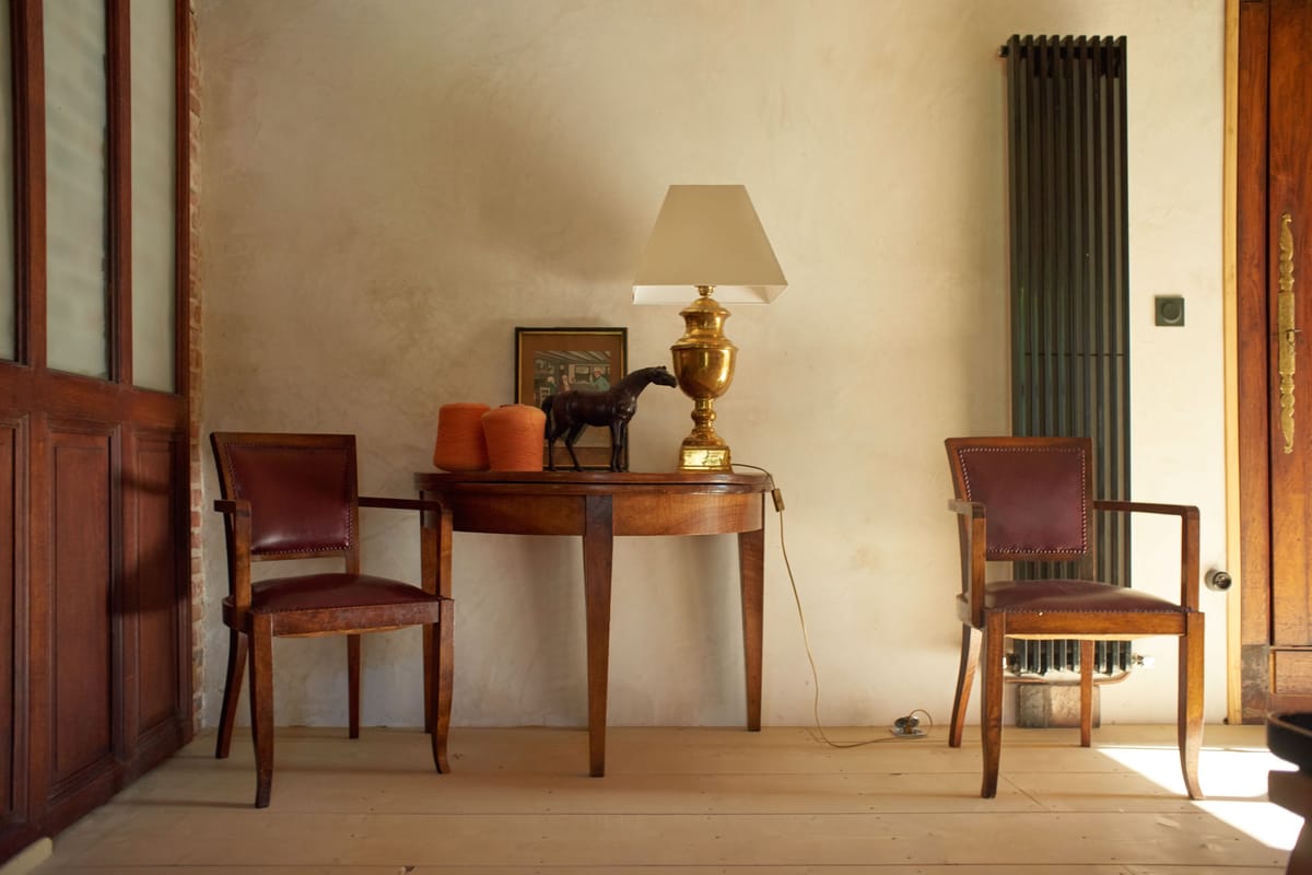 An interior detail shot of a room inside what appears to be a French château or manor house, showing no wedding activity or people. The scene features a demi-lune walnut console table flanked by two matching armchairs with dark burgundy leather upholstery and brass nail-head trim. On the console table sit two terracotta-orange cylindrical objects, a small dark bronze horse figurine, a small framed painting, and a brass urn-style table lamp with a cream square shade. The pale painted plank floor, bare plaster walls in off-white, a cast-iron vertical radiator in dark grey-green, and tall dark wood panelled doors contribute to a classic, worn French country interior aesthetic. Potential venue feature image.