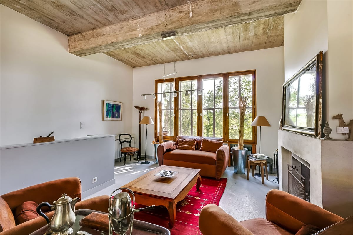 Interior wide shot of a living room space within what appears to be a French country property. The room features exposed wooden ceiling beams, white-painted walls, a white plaster fireplace with a large gilt-framed mirror above it, and a polished concrete floor. Seating consists of rust-orange upholstered armchairs and a cognac leather sofa arranged around a wooden coffee table set on a red patterned rug. A silver tea service is visible in the foreground. Large wood-framed French windows flood the room with natural light. Decor is mixed rustic and contemporary, with abstract artwork, a vintage black bistro chair, floor lamps with cream shades, and small sculptural objects on the mantelpiece. No people, wedding attire, or wedding-specific decor are present in this image. Potential venue feature image.