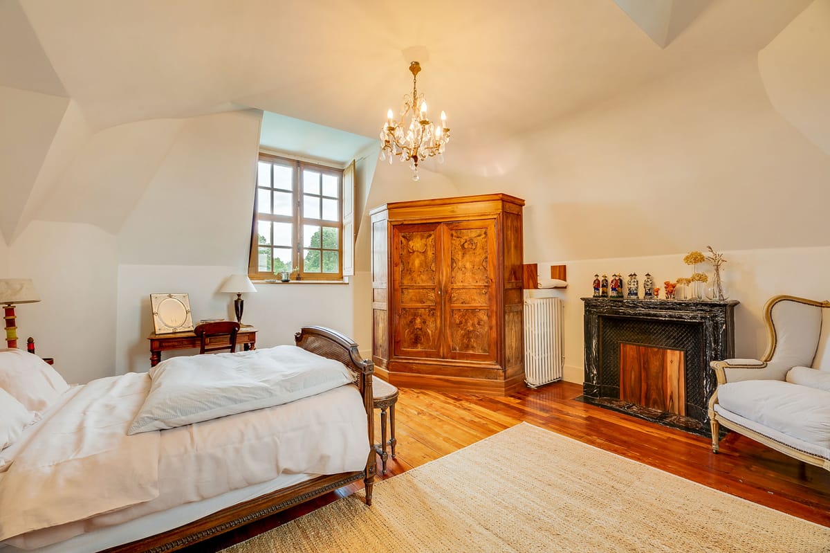 Interior wide shot of a château bedroom, likely used as a bridal suite or guest accommodation. The room features vaulted white ceilings, warm-toned hardwood flooring, and a natural fiber area rug. Furnishings include an antique carved wood bed dressed in white linens, a large burled walnut armoire, a dark mahogany bedside table with a table lamp, and a cream upholstered wingback chair in a French Louis XVI style. A black marble fireplace mantel is decorated with decorative ceramic figurines and a vase of dried ivory stems. A small brass and crystal chandelier hangs from the ceiling, and natural light enters through a multi-pane casement window. No people are present. Potential venue feature image.