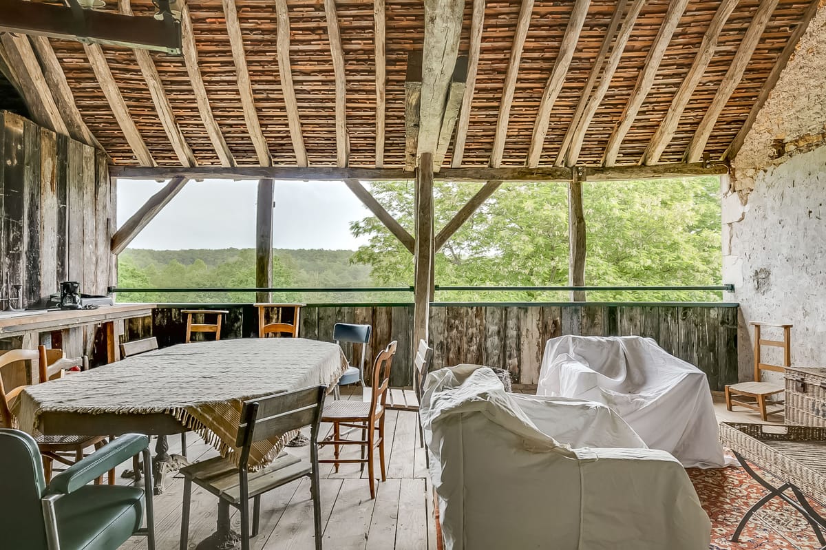 This image shows the interior of a rustic open-sided barn or agricultural outbuilding, not a wedding photograph. The space features exposed wooden beam rafters with terracotta roof tiles, weathered wooden plank walls and flooring, and a mix of mismatched wooden chairs surrounding a table covered with a fringed lace tablecloth. Several pieces of furniture are covered with white dust sheets. The structure opens onto a wide view of wooded countryside. The space appears to be in an undecorated, utilitarian state with no wedding styling, florals, or event setup visible. This image does not depict a wedding event or any wedding-related activity.