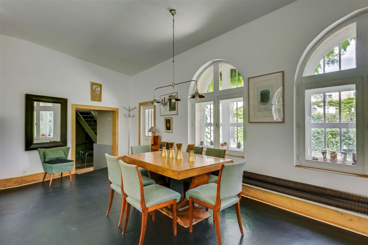 Wide-angle interior shot of a dining room inside what appears to be a French property venue. The room features a large rectangular wooden dining table surrounded by six mid-century style chairs upholstered in sage green houndstooth fabric, with brass hourglass-shaped decorative objects arranged down the center of the table. An industrial-style pendant light with multiple metal shades hangs above the table. The room has high white ceilings, dark painted floors, arched windows with white frames letting in natural light, and framed artwork on the walls. A green-framed mirror and a sage green upholstered armchair are visible in the background left corner. The overall aesthetic is mid-century modern with a muted green and warm wood palette. No people are present. Potential venue feature image.