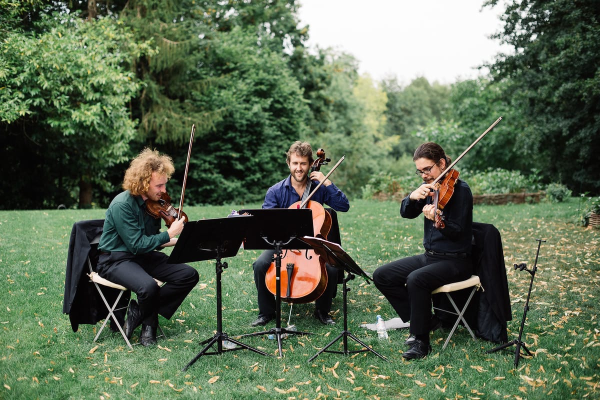 A string trio performs outdoors during what appears to be a wedding cocktail hour or ceremony, seated on folding chairs on a lawn with scattered fallen leaves. The three male musicians are playing two violins and a cello, with black music stands positioned in front of them. The musician on the left wears a dark green shirt with black trousers, the cellist in the center wears a navy blue shirt, and the violinist on the right is dressed entirely in black. All three are actively playing, with the cellist smiling toward the camera. The setting is a garden or parkland with dense mature trees in the background. Wide shot, taken at ground level.
