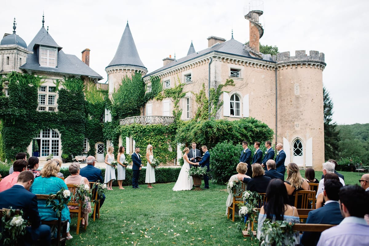An outdoor wedding ceremony taking place on a lawn directly in front of a large French château featuring turrets, slate-roof towers, ivy-covered stone façades, and Gothic-arched windows. The bride, in a white gown, and groom, in a navy suit, stand facing an officiant who holds an open book, with the couple appearing to exchange vows or rings. Two bridesmaids in light grey midi dresses hold small bouquets of white flowers and greenery, while approximately four groomsmen in navy suits stand on the opposite side. Aisle chairs in natural wood are decorated with white floral and greenery arrangements, and potted arrangements with lush greenery flank the ceremony area. Approximately 40–50 seated guests are visible on both sides of the aisle, dressed in smart-casual attire in a range of colors. The overall decor palette is white and green with a classic, understated style. Wide-angle shot taken from behind and slightly elevated above the seated guests. Potential venue feature image.