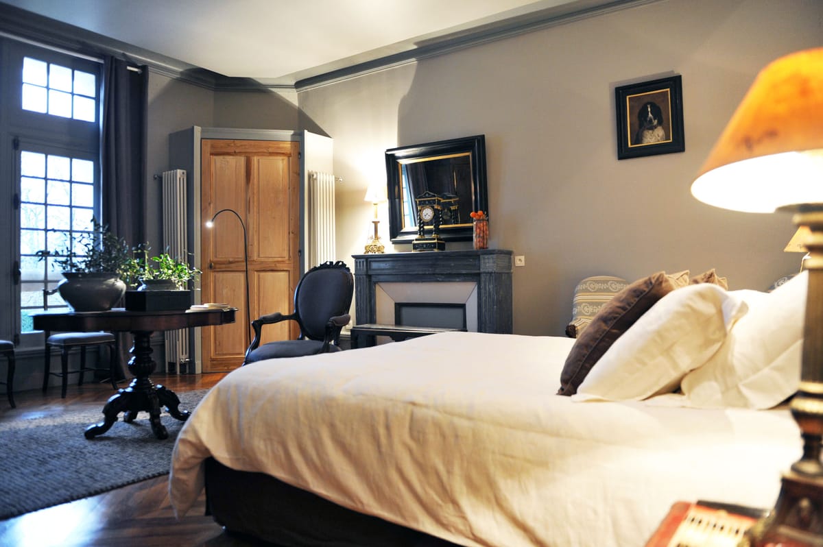 Interior shot of a guest bedroom, likely within a château or manor house venue, decorated in a classic French style. The room features a double bed dressed in white linen with ivory and dark brown accent pillows, a grey marble fireplace mantel topped with a black antique clock and a gilt candelabra lamp, and a large black-framed mirror. Furnishings include a dark wood pedestal table with potted green plants, a dark blue Louis XVI-style armchair, and a brass bedside lamp with an amber shade. The walls are painted in a warm taupe-grey with white crown molding, the floor is dark hardwood with a grey area rug, and tall multi-pane windows with charcoal curtains allow natural light into the space. Potential venue feature image.
