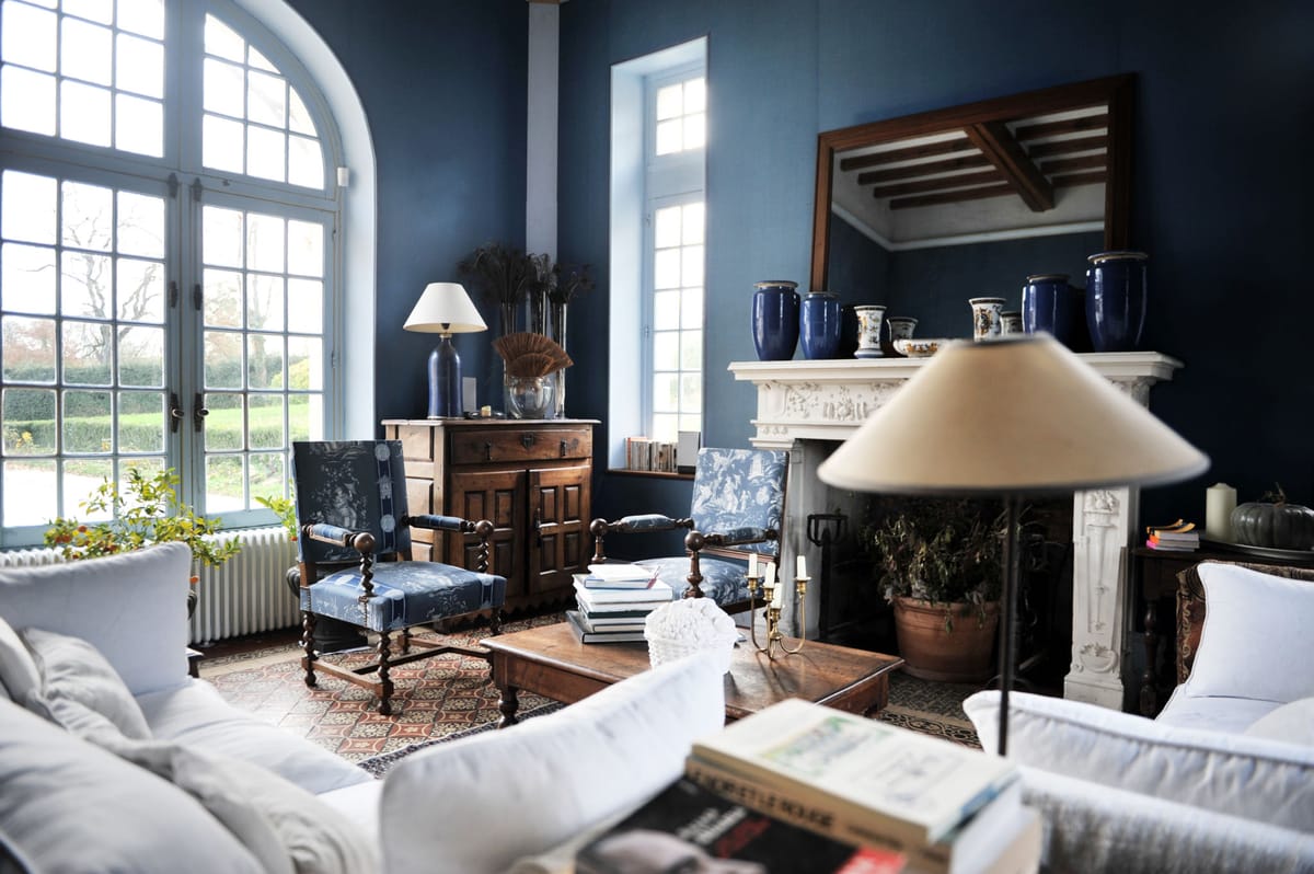 Interior wide shot of a formal salon or sitting room inside what appears to be a French château or manor house. The room features deep navy blue walls, a carved white marble fireplace mantel decorated with cobalt blue ceramic vases and white-and-blue faience pottery, and two antique armchairs upholstered in blue toile de Jouy fabric with barley-twist wooden legs. White linen sofas and a large floor lamp with a cream shade are positioned in the foreground, while a dark wood antique cabinet and table lamp with a white shade sit near tall arched French windows overlooking formal hedged grounds. A patterned encaustic tile floor is partially covered by a geometric rug, and a large wood-framed mirror hangs above the fireplace. No people are present. Potential venue feature image.