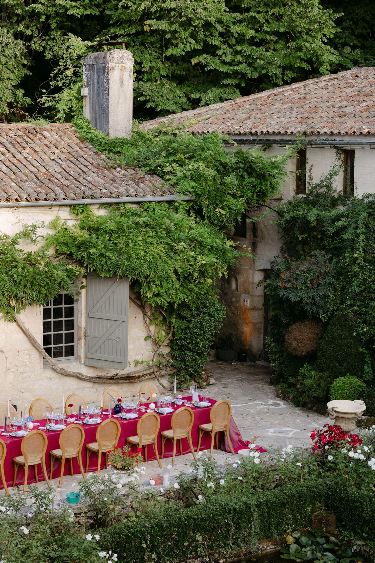 An outdoor reception dining setup photographed from an elevated wide-angle perspective in a French countryside courtyard. A long rectangular table is dressed in a deep magenta/fuchsia linen tablecloth and set with glassware, white plates, and scattered candles in cobalt blue and taper styles, surrounded by approximately ten natural wood cane-back medallion chairs on the visible side. The setting is a rustic stone-walled property with terracotta-tiled roofs, grey-green shuttered windows, and climbing vines covering much of the facade, styled in a rich, jewel-toned color palette that contrasts with the aged stone architecture. White and red flowering plants are visible in the foreground and near a stone planter to the right. Potential venue feature image.