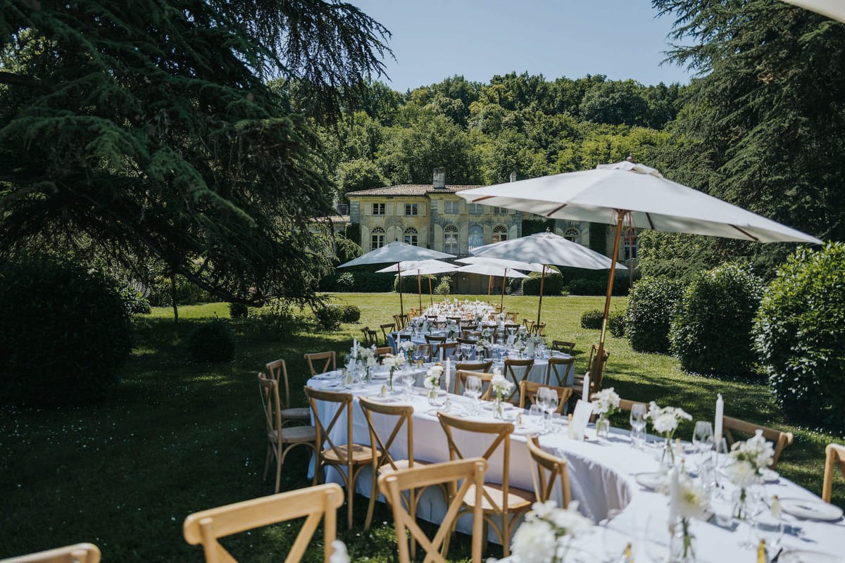 An outdoor wedding reception setup on the grounds of a French manor house, photographed from a slightly elevated wide-angle perspective. Multiple dining tables — a mix of long rectangular and round — are dressed in white linen and set with wine glasses, white taper candles, and small bud vases of white flowers, arranged across a lawn with natural wood cross-back chairs. Large white market umbrellas on copper-toned poles are positioned throughout the setup to provide shade. The decor palette is white and natural wood with a classic, understated style. In the background, a two-storey stone manor house with arched windows and a tiled roof is visible, surrounded by mature trees and manicured hedging. No guests are present — the space is fully set and ready for service. Potential venue feature image.