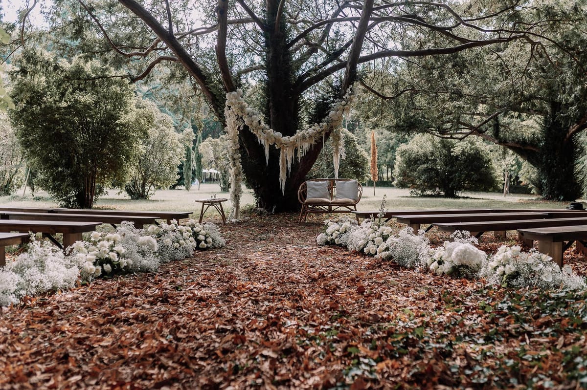 An outdoor ceremony setup photographed before the ceremony, with no people present. The altar is centered beneath a large tree, with a floral garland of white roses and baby's breath draped in a crescent arc across the trunk, accented with hanging macramé or fringe details in a boho style. A rattan loveseat with white cushions serves as the couple's seat, flanked by a small wooden side table. Dark wood benches are arranged in two rows forming an aisle, which is lined on both sides with loose clusters of white hydrangeas, white roses, and baby's breath arranged directly on the ground. The aisle floor is covered in rust-colored fallen leaves. The overall decor palette is all-white florals against natural wood tones, consistent with a rustic-boho aesthetic. Wide establishing shot taken from the back of the aisle looking toward the altar. Potential venue feature image.