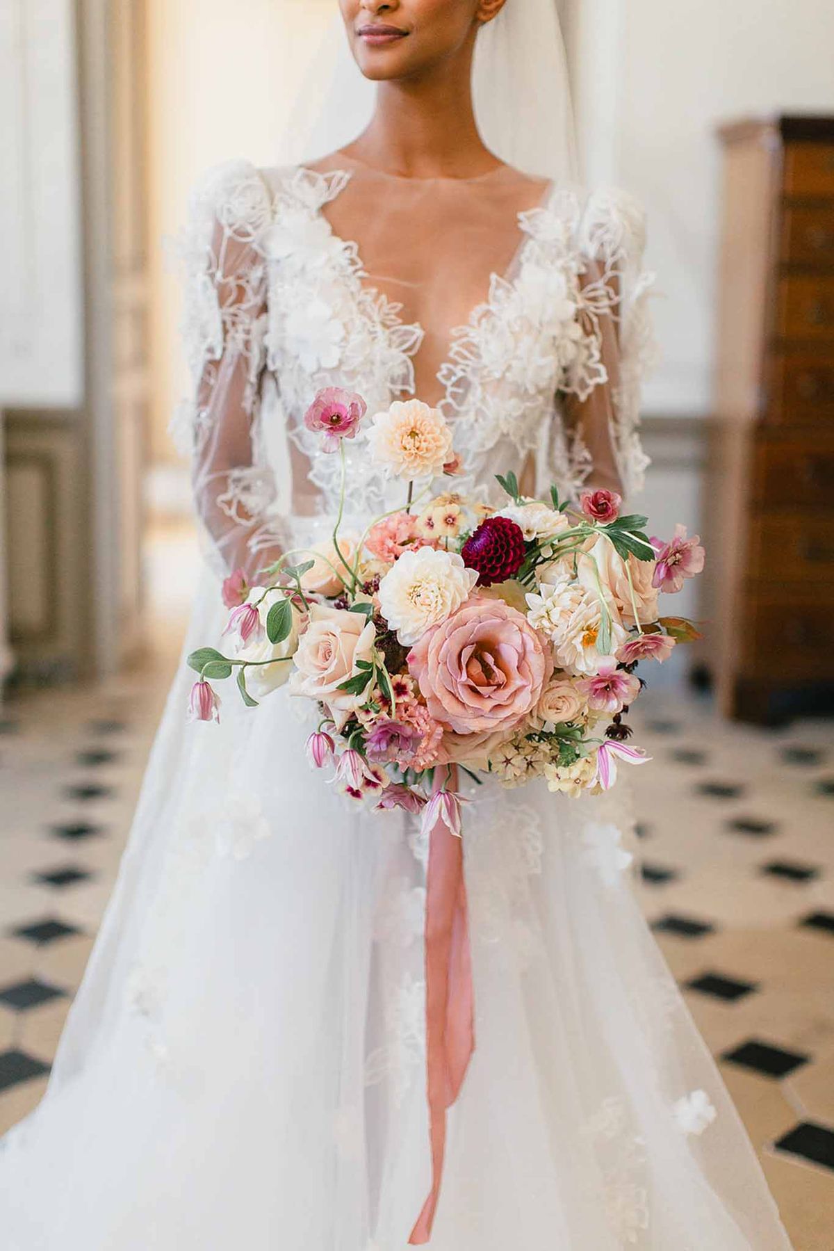 A close-up portrait of a bride standing indoors, cropped from the chin to mid-skirt, holding a large garden-style bouquet. She wears a white A-line gown with a deep V-neckline, sheer long sleeves, and three-dimensional floral lace appliqué across the bodice. The bouquet features blush and dusty rose garden roses, peach dahlias, burgundy dahlias, ivory peonies, pink poppies, hanging clematis, and small filler blooms in cream and mauve, tied with a long trailing dusty rose silk ribbon. The setting is a classical interior with black-and-white diamond-patterned marble tile flooring and pale panelled walls, suggesting a château or manor house.