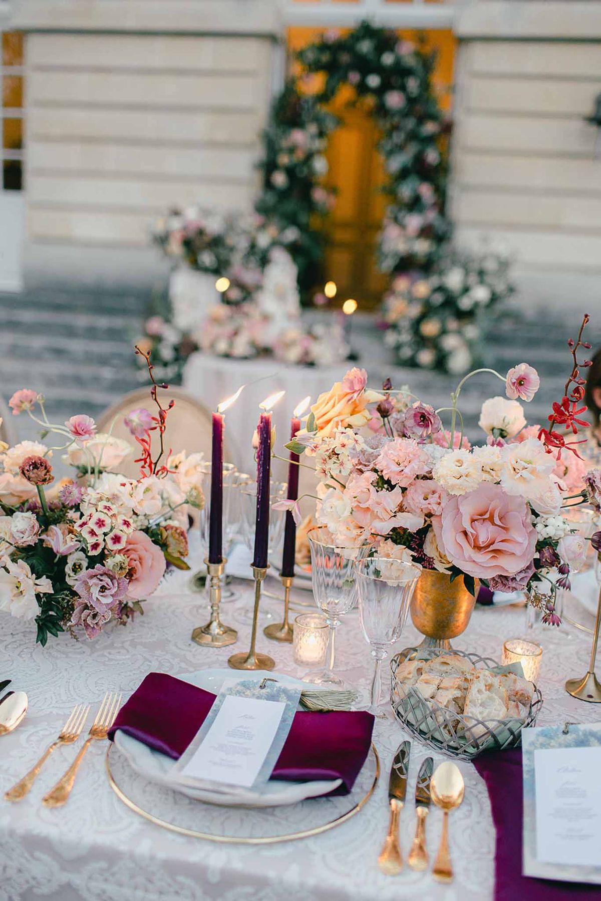A close-up detail shot of an outdoor wedding reception table set in front of a classical building entrance, with a floral arch visible over the doorway in the background. The table is dressed in a white jacquard linen and styled with a rich jewel-toned and blush palette: deep plum satin napkins, gold flatware, gold-rimmed white charger plates, and faceted crystal glassware. Floral centerpieces feature blush garden roses, cream ranunculus, mauve lisianthus, red orchid stems, and mixed blooms arranged in gold urn vases. Three tall deep purple taper candles in brass candlestick holders are lit, alongside small votive candles scattered across the table. A printed menu card is placed on each setting, and a wire basket holds bread rolls. In the soft-focus background, a tiered wedding cake is visible at the base of the steps, surrounded by additional loose floral arrangements in blush and white tones. The overall styling theme is classic and ornate, combining gold, plum, and blush tones.