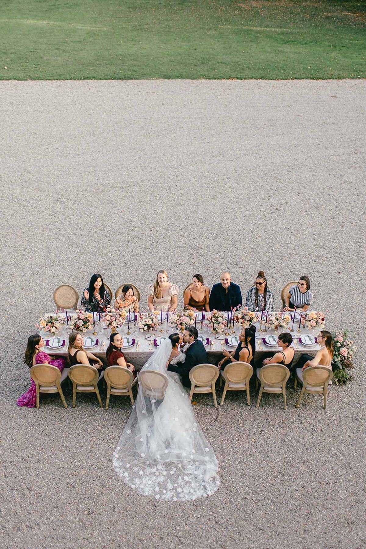An aerial shot of an intimate outdoor wedding reception dinner set on a gravel courtyard, with approximately 12 guests seated around a single long rectangular table on Louis XVI-style cane-back chairs. The bride and groom are seated at the center of the table sharing a kiss; the bride wears a white gown with a cathedral-length veil featuring floral appliqué scattered across the train, which fans out prominently across the gravel below her. The table is dressed with a light linen runner and styled with clusters of blush, peach, dusty rose, and burgundy blooms — likely including roses and ranunculus — arranged in low gold vessels running the full length of the table, interspersed with tall deep purple taper candles in gold candleholders. Place settings include white plates with purple chargers or napkins and gold flatware. Guests are dressed in a mix of colorful and dark attire including a fuchsia gown, burgundy top, bronze dress, and dark suits, giving the gathering a festive, eclectic feel. The overall styling palette combines jewel tones and warm neutrals in a modern, slightly maximalist approach.