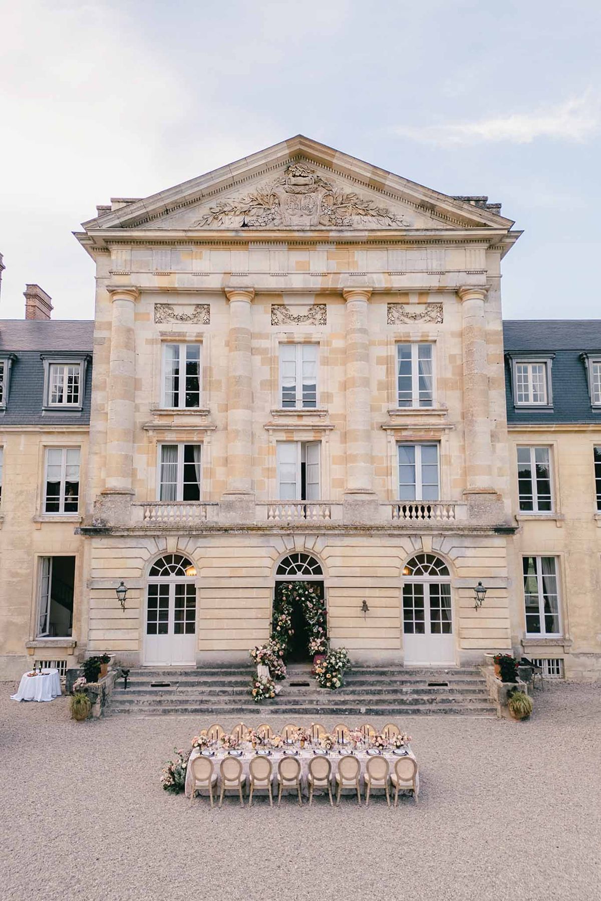 An outdoor wedding reception setup photographed from a wide-angle, straight-on perspective in front of a classical French château with cream-colored limestone architecture, a pediment with carved stonework, and arched ground-floor windows. A long rectangular dining table seating approximately 14 guests is positioned on the gravel forecourt directly in front of the château's main entrance steps, set with dark taper candles, floral centerpieces in soft pink and peach tones, and what appear to be dark charger plates. French-style medallion-back chairs in a natural/greige finish flank both sides of the table. The central doorway of the château is framed by a large, lush floral installation featuring pink, peach, and cream blooms with trailing greenery, and additional floral arrangements are placed at the base of the entrance steps. The overall decor palette combines blush, peach, and cream florals with warm candlelight against the pale stone facade. Potential venue feature image.