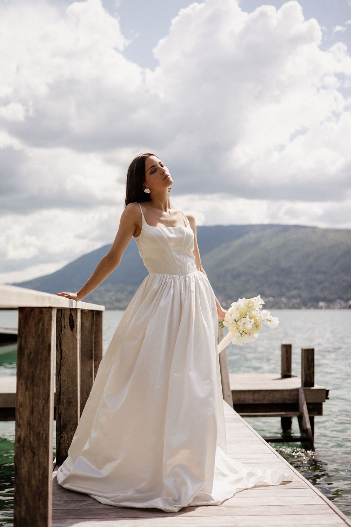 A bridal portrait taken outdoors on a wooden lakeside dock, with a mountain lake and rolling hills visible in the background. The bride, a dark-haired woman, stands alone in a full-length white gown with thin spaghetti straps, a fitted bodice, and a full gathered skirt with a short train; she wears statement gold floral drop earrings and holds a compact bouquet of white sweet peas or ranunculus loosely at her side. Her head is tilted upward with eyes closed in a posed, editorial style. The composition is a medium-full portrait shot taken at slight low angle, giving prominence to the dress silhouette against the open water and sky backdrop, in a clean, modern bridal style.