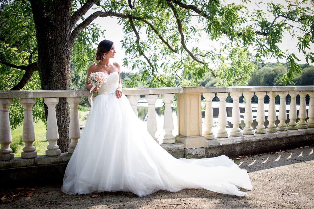 A bridal portrait taken outdoors on a stone balustrade terrace overlooking a lake or river, with a classic French château or estate setting. The bride stands alone, gazing to one side, wearing a white strapless ball gown with a lace sweetheart bodice, full tulle skirt, and an extended cathedral-length train spread across the terrace floor. She holds a rounded bouquet of blush pink roses and small white filler flowers tied with a white ribbon, and wears her dark hair in an updo with drop earrings. The composition is a full-length portrait shot framing the bride against the stone balustrade, with water visible in the background. The styling is classic and formal.