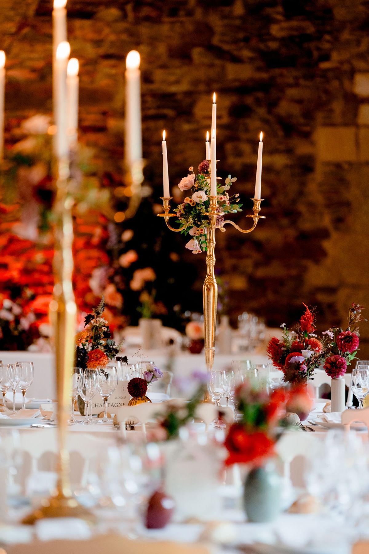 A close-up detail shot of a wedding reception table setup inside a stone-walled venue, likely a barn or château outbuilding. The centerpiece is a tall gold candelabra holding five lit white taper candles, adorned at its base with a small floral arrangement of blush roses, burgundy dahlias, and green foliage. The tables are dressed in white linen and set with stemmed wine glasses, white plates, and silverware, with a visible card reading 'CHAMPAGNE.' Scattered along the table are small bud vases in sage green, amber, and burgundy tones, each holding clusters of deep red dahlias, orange blooms, blush roses, and purple wildflowers, creating a rich, jewel-toned autumn palette. In the soft background, a large floral installation in red, orange, and pink tones is visible against the exposed stone wall, and additional tall gold taper candelabras frame the foreground.