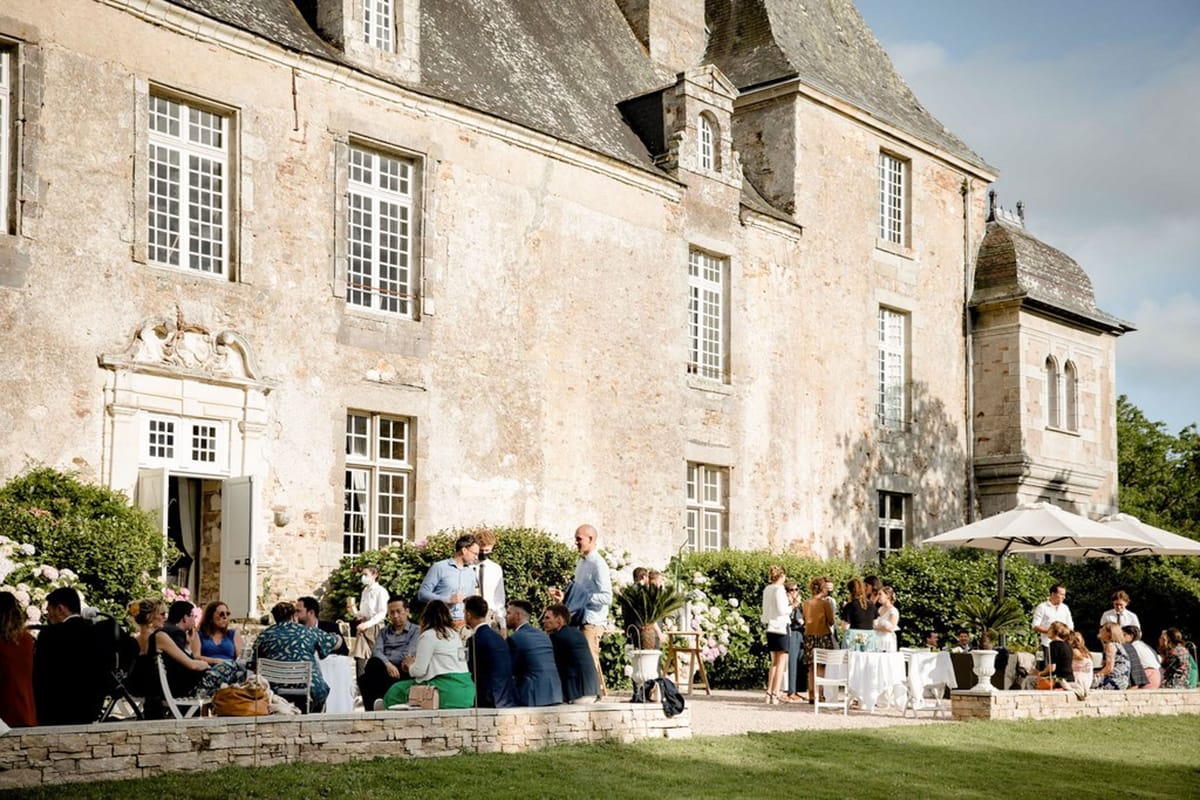 An outdoor cocktail hour taking place on the terrace directly in front of a French château, with approximately 30–40 guests mingling, seated on white folding chairs, and standing in small groups. The château is a large multi-story stone building with a steep slate roof, ornate white carved doorframe, and tall white-framed windows. White cocktail tables with white linen cloths and large white urn planters are positioned along the terrace, with cream parasols providing shade on the right side. Guests are dressed in smart-casual attire including navy suits, a green skirt, blue dresses, and light summer shirts. White flowering hydrangea bushes line the base of the château façade, adding decorative interest along the terrace. The shot is a wide environmental image taken from the lawn, capturing both the full venue façade and the social activity in front of it in bright afternoon sunlight. Potential venue feature image.