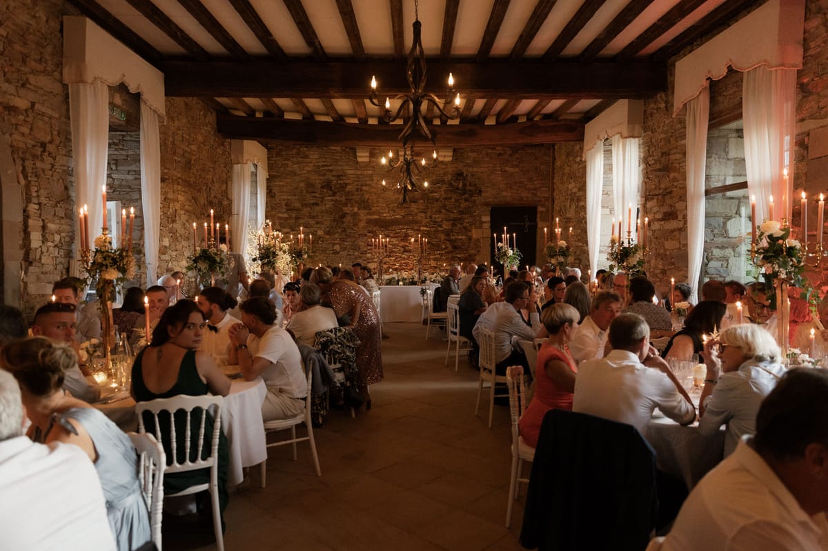 A wedding reception dinner is underway in a rustic indoor hall with exposed stone walls, dark wooden beam ceilings, and a wrought-iron chandelier with Edison-style bulbs hanging centrally. Approximately 60–80 guests are seated at round and rectangular tables arranged in two rows along the length of the room, with a head table visible at the far end. White Chiavari chairs are used throughout. The decor palette is warm and candlelit, with tall terracotta-toned taper candles on gold candelabras and floral arrangements featuring white blooms and greenery placed on each table. White sheer curtains frame the stone archway windows on both sides of the room. The overall styling is classic French rustic with warm amber lighting from candles and the chandelier creating a golden tone across the space. Wide shot taken from the entrance end of the room looking toward the head table.