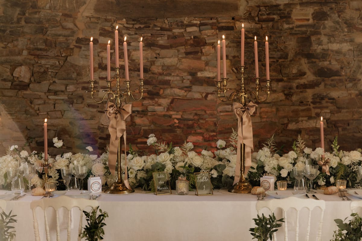 A detail shot of a wedding reception sweetheart table set against an exposed stone wall interior, with a table sign reading 'Table des Mariés' confirming it as the couple's table. Two tall brass candelabras, each holding five lit dusty rose taper candles and tied with large champagne satin ribbon bows, flank a low runner of ivory roses, white carnations, gypsophila, and mixed greenery that spans the full length of the table. The white linen-covered table is set with ribbed glassware, silver cutlery, small gold votive candles, individual bread rolls, and printed place cards or menu cards. White Chiavari-style chairs are visible in the foreground, and a green card table number marker labeled 'Table 1' sits alongside the bridal table sign. The overall decor palette is ivory, champagne, and dusty rose with gold metallic accents, consistent with a classic romantic styling theme.
