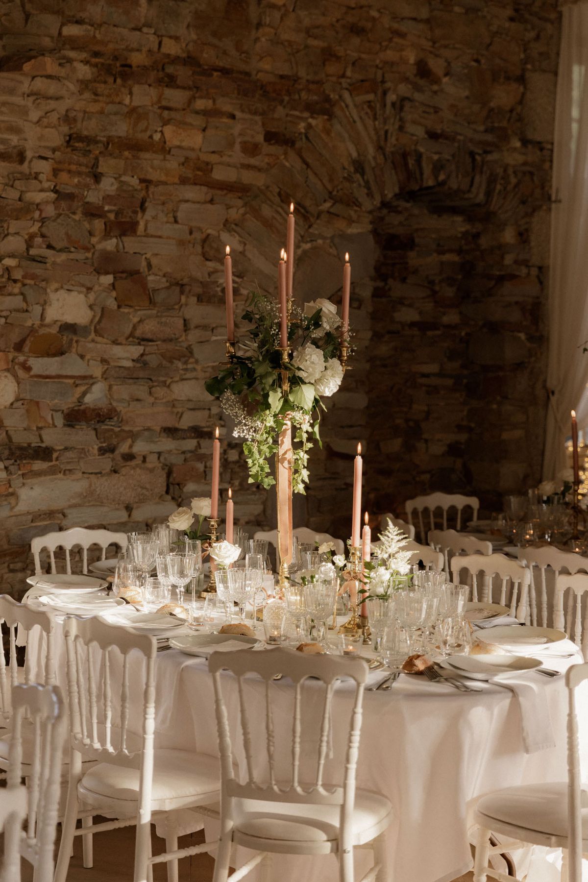 A reception table detail shot taken indoors in a venue with exposed stone walls and an arched architectural feature. The round table is dressed in a white linen tablecloth and surrounded by white Napoléon-style chairs. The centerpiece is a tall gold candelabra holding five lit dusty rose taper candles, topped with an arrangement of white hydrangeas, white roses, baby's breath, and trailing green ivy. Additional shorter gold candleholders with matching dusty rose tapers are placed along the table, alongside low white rose and fern accents. The table is set with white plates, white linen napkins, silver cutlery, and multiple crystal glasses including wine and tumbler styles. Small bread rolls are visible at each place setting. The overall decor palette is white, gold, and dusty rose with greenery, in a classic French style. Additional similarly dressed tables are visible in the background.