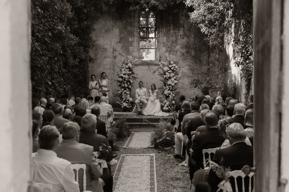 This black-and-white wide shot captures a wedding ceremony taking place inside the roofless ruins of a chapel or abbey, with ivy-covered stone walls and a Gothic arched window visible in the background. The couple is seated on a raised platform at the altar end of the space, framed by two large floral columns featuring full blooms and foliage in light tones, with fern accents placed along the base of the platform. An officiant stands at a podium to the left, reading from notes, while approximately 80–100 guests are seated on white chairs arranged in two sections separated by a central aisle lined with patterned rugs. The groom wears a light-colored suit and the bride wears a full-length white gown and holds a bouquet. The image is shot from the doorway looking inward, giving a framed, documentary perspective with strong contrast between the bright altar area and the darker tonal foreground of the seated guests. Potential venue feature image.