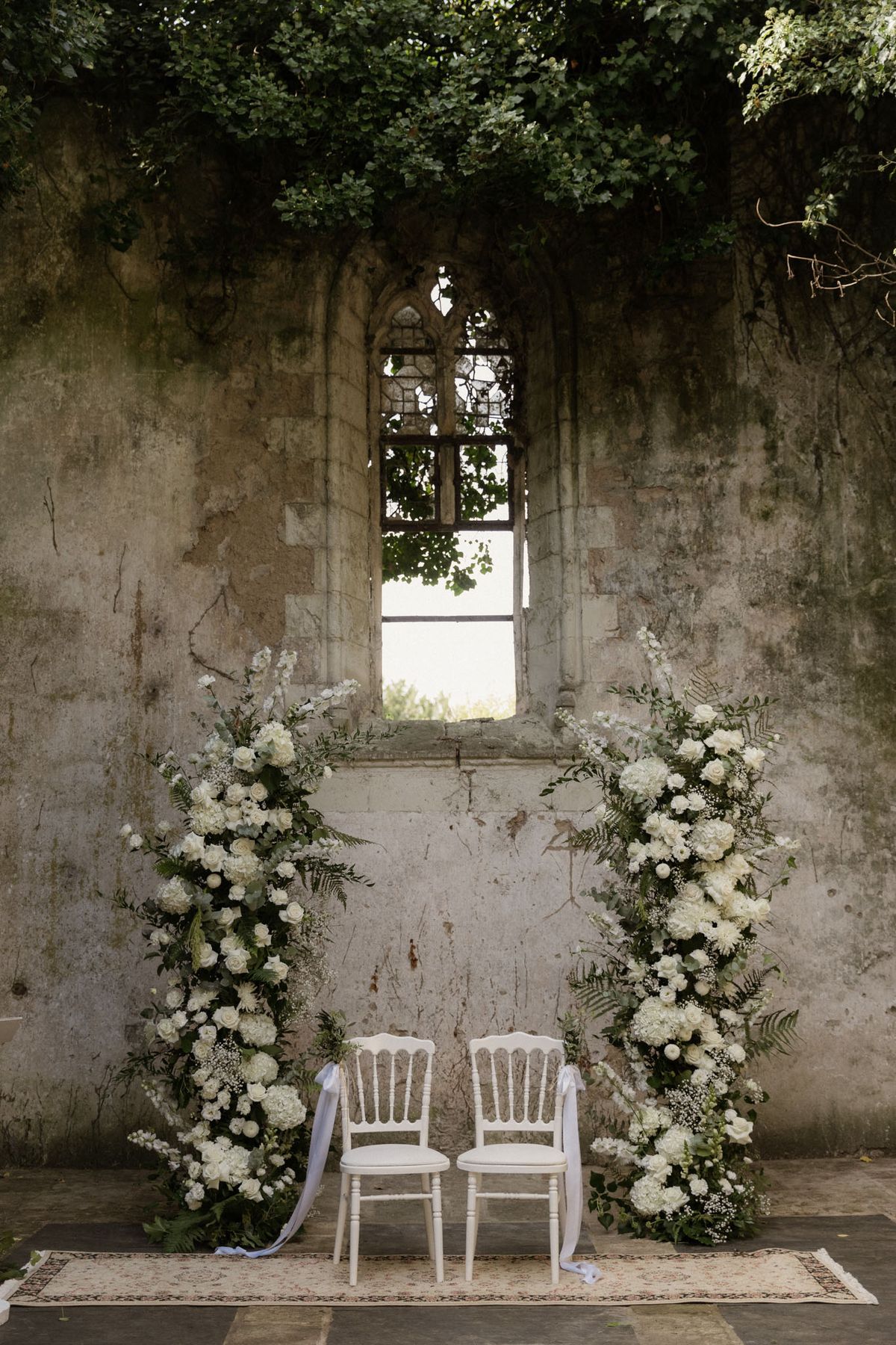 An outdoor ceremony setup photographed against the weathered stone wall of a roofless ruined chapel or historic structure, with a Gothic arched window as the central backdrop. Two white Chiavari-style chairs are placed side by side on a patterned vintage rug, flanked by two tall freestanding floral columns composed of white hydrangeas, white roses, baby's breath, ferns, and trailing greenery. Soft white ribbon or fabric drapes loosely from each chair. The overall color palette is white and deep green, creating a classic, nature-forward aesthetic. Wide shot, no people present. Potential venue feature image.