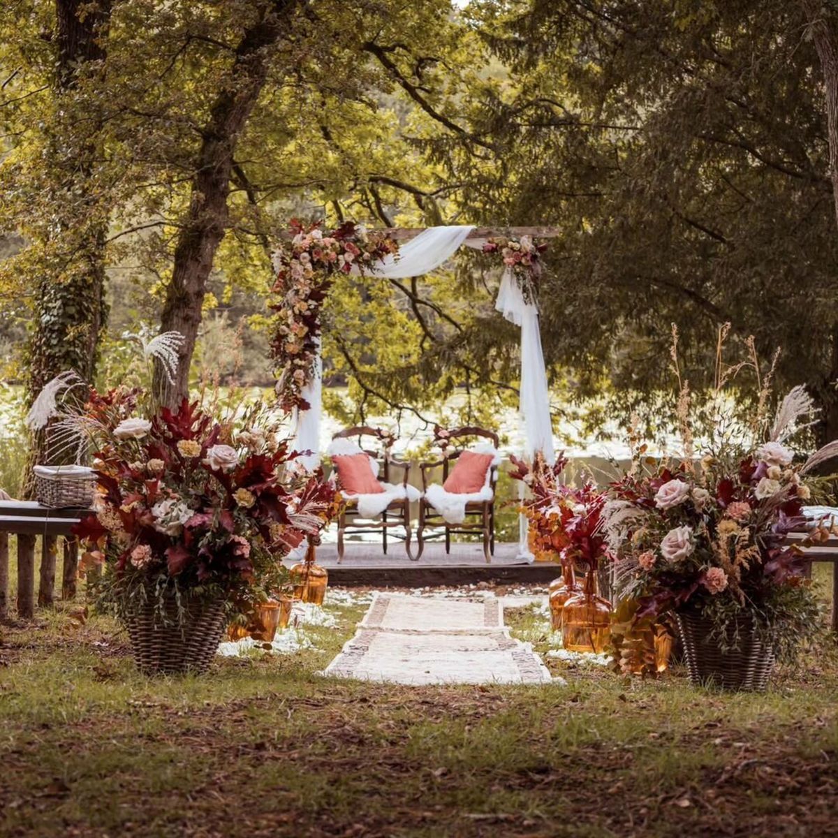 An outdoor ceremony setup photographed from the aisle entrance, with no people present. The ceremony space is set beneath mature trees with a body of water visible in the background. A white draped wooden arch serves as the focal point, decorated with abundant florals in deep burgundy, blush pink, rust, and cream tones including roses, dried tropical leaves, and pampas grass. Two dark wood antique chairs with coral-orange cushions and white sheepskin throws are positioned beneath the arch on a raised platform. The aisle runner is a neutral-toned vintage-style rug flanked by large wicker basket arrangements overflowing with the same rich autumn-palette florals, and amber glass vase lanterns line the path. Wooden pew-style benches are partially visible on either side. The overall styling is boho-rustic with a warm autumnal color palette. Wide shot taken from ground level looking toward the arch.