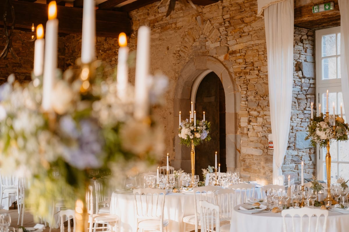 Indoor wedding reception setup inside a historic stone-walled venue with exposed wooden ceiling beams and a distinctive Gothic-arched alcove as a backdrop. Round tables are dressed in white linen and set with crystal glassware, white plates, and scattered gold accents, surrounded by white Louis ghost-style chairs. Tall gold candelabras hold white taper candles and are topped with lush floral arrangements featuring white blooms, soft lavender flowers, and greenery including eucalyptus. White sheer draping frames a window on the right side of the frame, and warm candlelight fills the room. The foreground features a softly blurred candelabra and floral arrangement, giving depth to the wide interior shot. The overall decor palette combines white, gold, and muted blue-lavender tones in a classic, warm style. Potential venue feature image.