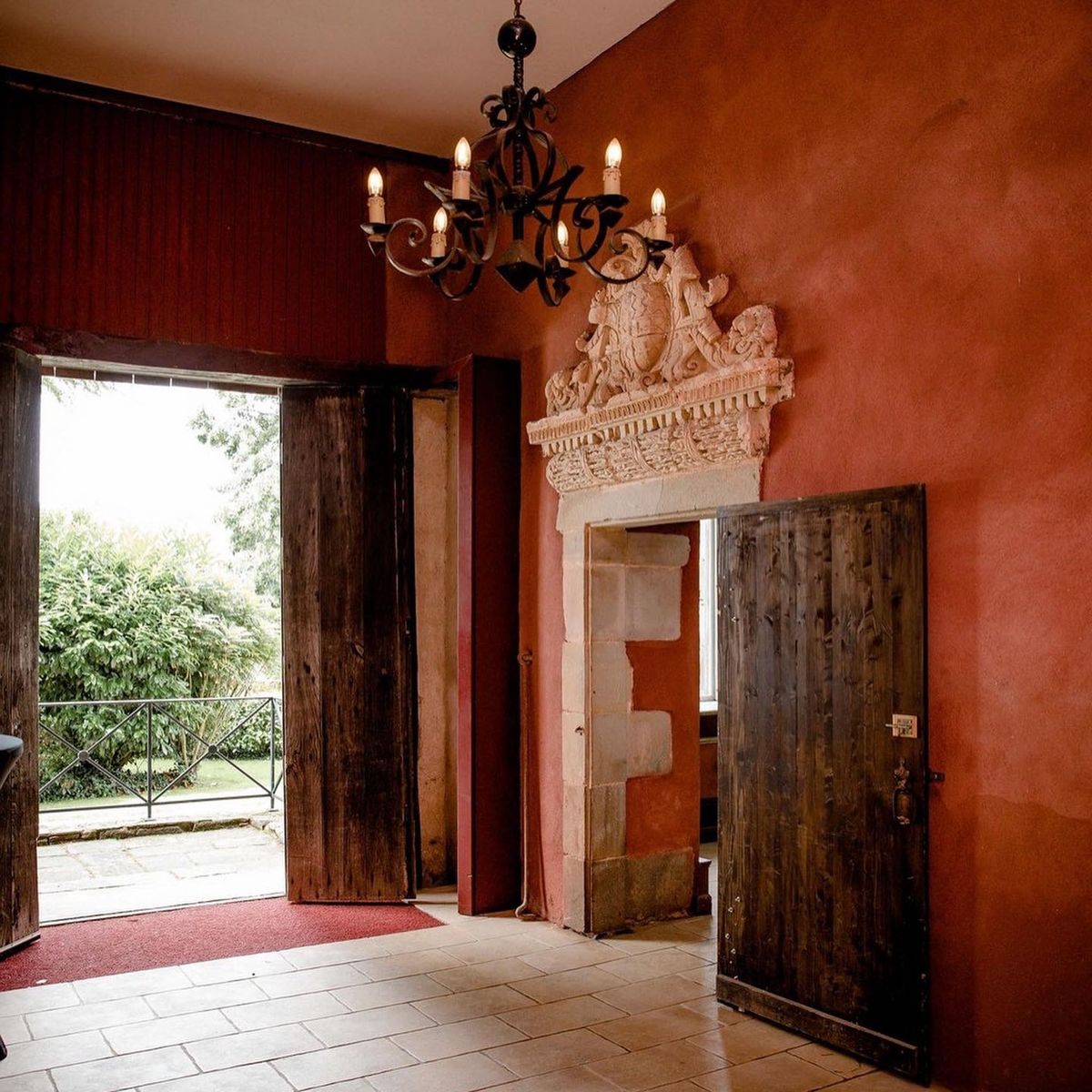 An interior architectural shot of a historic venue entrance hall featuring deep terracotta-red plastered walls and large-format stone tile flooring. A wrought-iron candelabra-style chandelier with candle-bulb lights hangs from the white ceiling. Two doorways are visible: a wide open doorway with aged dark wooden doors leading to an exterior garden with a wrought-iron balustrade, fitted with a red floor runner at the threshold, and a second doorway topped with an elaborate carved stone pediment featuring a coat of arms with heraldic figures in cream-white stone. The heavy dark wooden door of the second opening is ajar, revealing a glimpse of a window beyond. The overall interior style is rustic historic, with raw aged materials and no people present. Wide interior shot. Potential venue feature image.