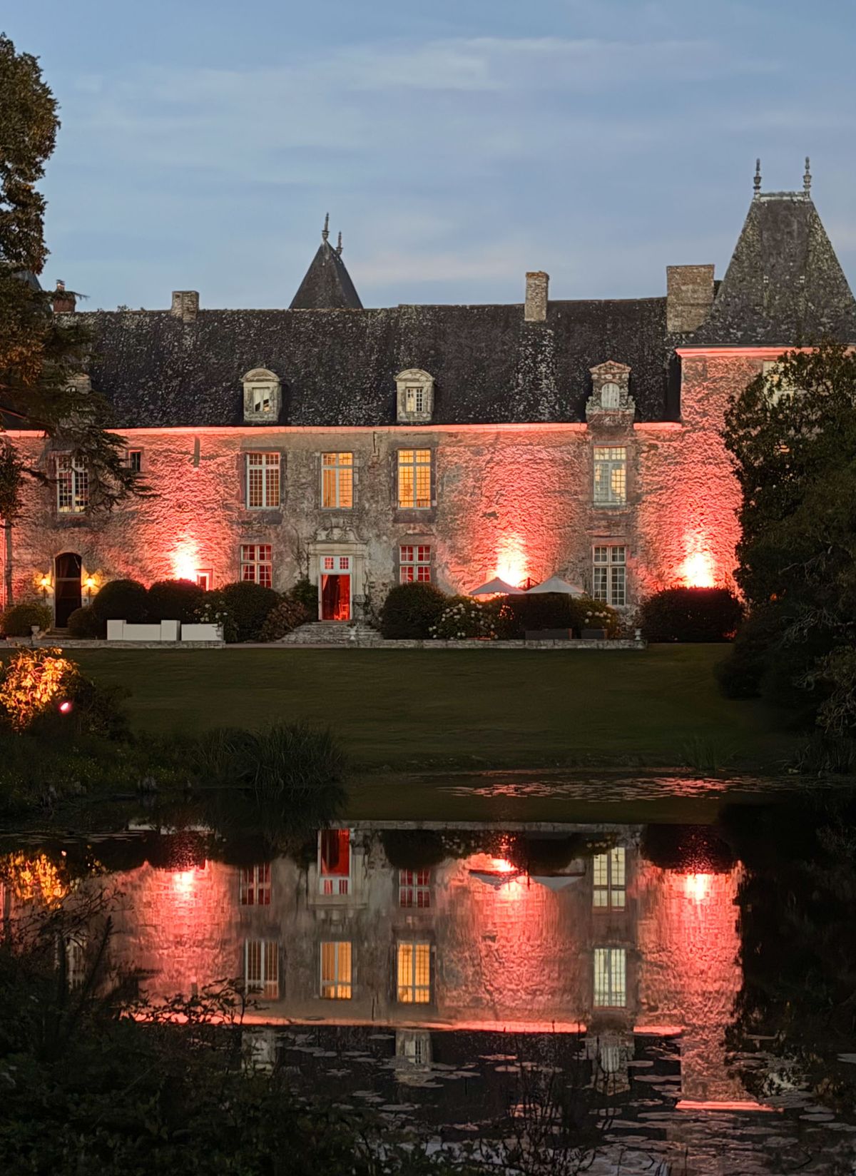 A wide exterior shot of a French château at dusk, photographed from across an ornamental pond. The stone manor house features a slate mansard roof with pointed dormers and decorative finials, multiple sets of tall French windows, and a central entrance with steps leading to a red door. The façade is lit with warm pink-red uplighting at ground level, while the interior windows glow amber, and the full reflection of the building and its lighting is visible in the still water of the pond in the foreground. White event parasols or canopy structures are visible on the lawn to the right of the entrance, suggesting an outdoor reception or cocktail hour is underway. Potential venue feature image.