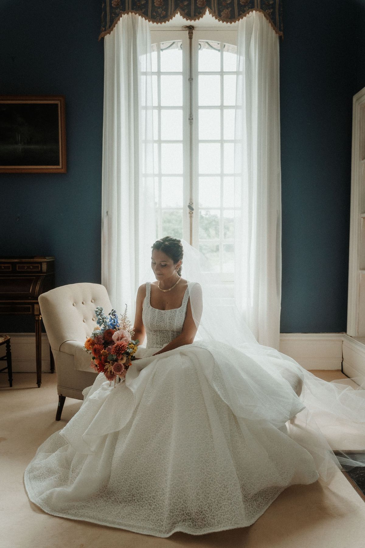 A bridal portrait taken indoors in a room with deep teal-blue painted walls, ornate curtain valances, and a tall arched French window with sheer white drapes providing soft backlight. The bride is seated on the floor in front of a cream button-back armchair, wearing a full ball gown with a square neckline, lace or textured fabric bodice, and a voluminous layered skirt with a long train spread across the floor. She wears a pearl necklace, drop earrings, and a veil, with her dark hair styled in an updo. She looks downward at a richly colored bouquet featuring burnt orange and rust dahlias, blush pink blooms, blue delphiniums, and terracotta-toned foliage. A gilt-framed painting and a dark antique writing desk are visible on the wall to the left, suggesting a château interior. Full-length portrait composition with natural window light.