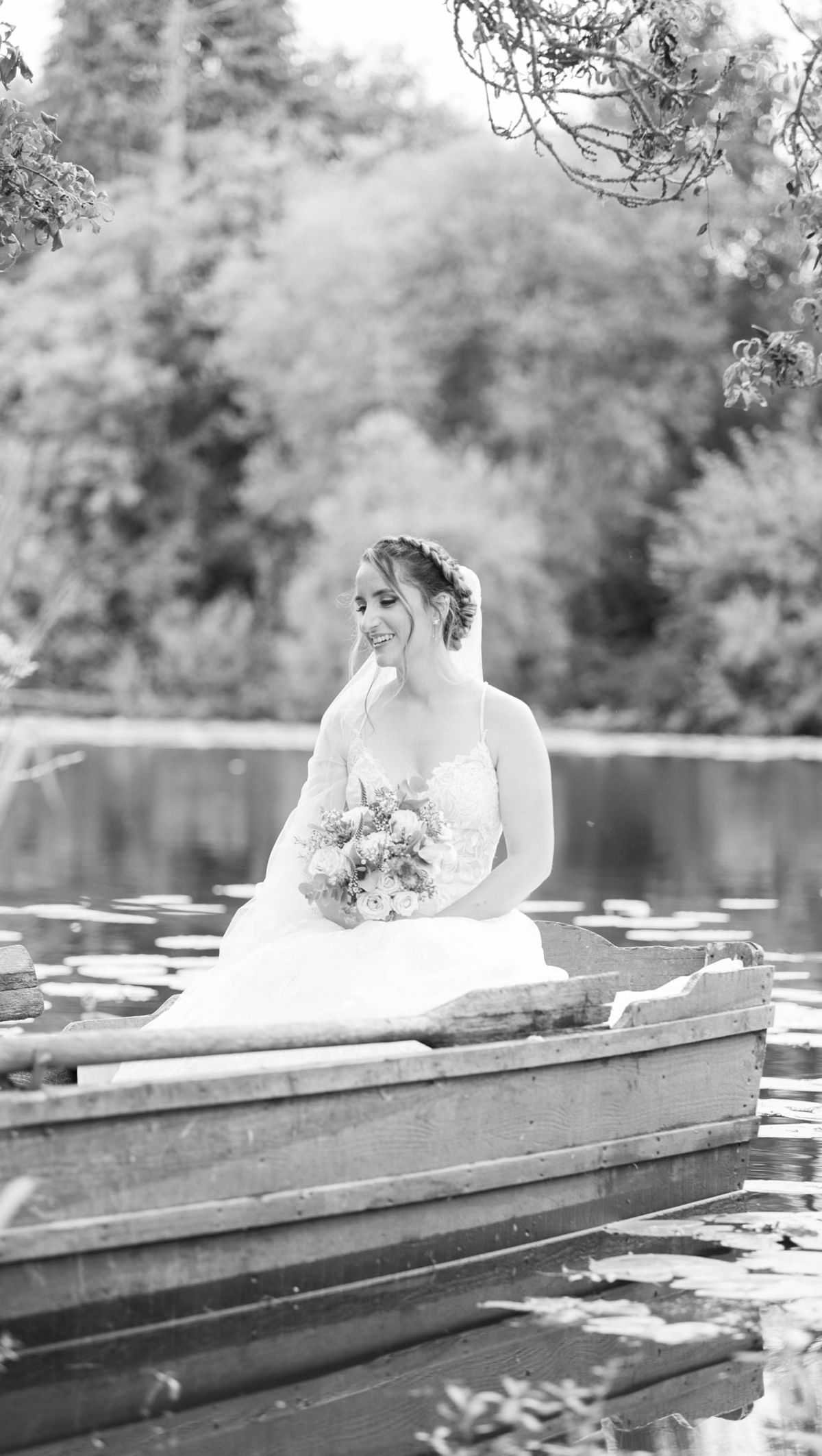 This is a black-and-white bridal portrait of a single bride seated in a wooden rowing boat on a pond with lily pads visible on the water's surface. The bride wears a full-skirted A-line gown with a lace bodice and thin straps, and her hair is styled in an updo with a braided crown detail; a veil drapes behind her. She holds a rounded bouquet featuring roses and what appear to be wildflower-style blooms with textured foliage, rendered in soft mid-tones against the bright white of her dress. The image is shot at a medium portrait distance with a shallow depth of field, placing the bride in sharp focus while the background dissolves into soft tones, and the overall contrast is light and airy with bright highlights and gentle shadows.