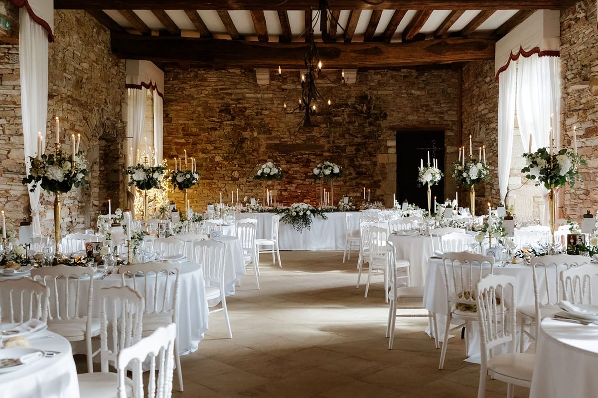 A wide shot of a wedding reception room set up before guests arrive, located inside a rustic château or manor hall with exposed stone walls, wooden beam ceiling, and stone tile flooring. The room features multiple round guest tables dressed in white floor-length linen with white Napoleon chairs, centered around a long rectangular head table also draped in white. Tall gold candelabras hold slender white taper candles and are topped with large arrangements of white roses, white hydrangeas, and trailing eucalyptus foliage. Lower floral garlands of greenery and white blooms run along the head table. A wrought-iron chandelier with Edison-style bulbs hangs from the ceiling at center, and sheer white curtains with burgundy valances frame the windows. The overall decor palette is white and gold with green accents, styled in a classic formal aesthetic. Potential venue feature image.
