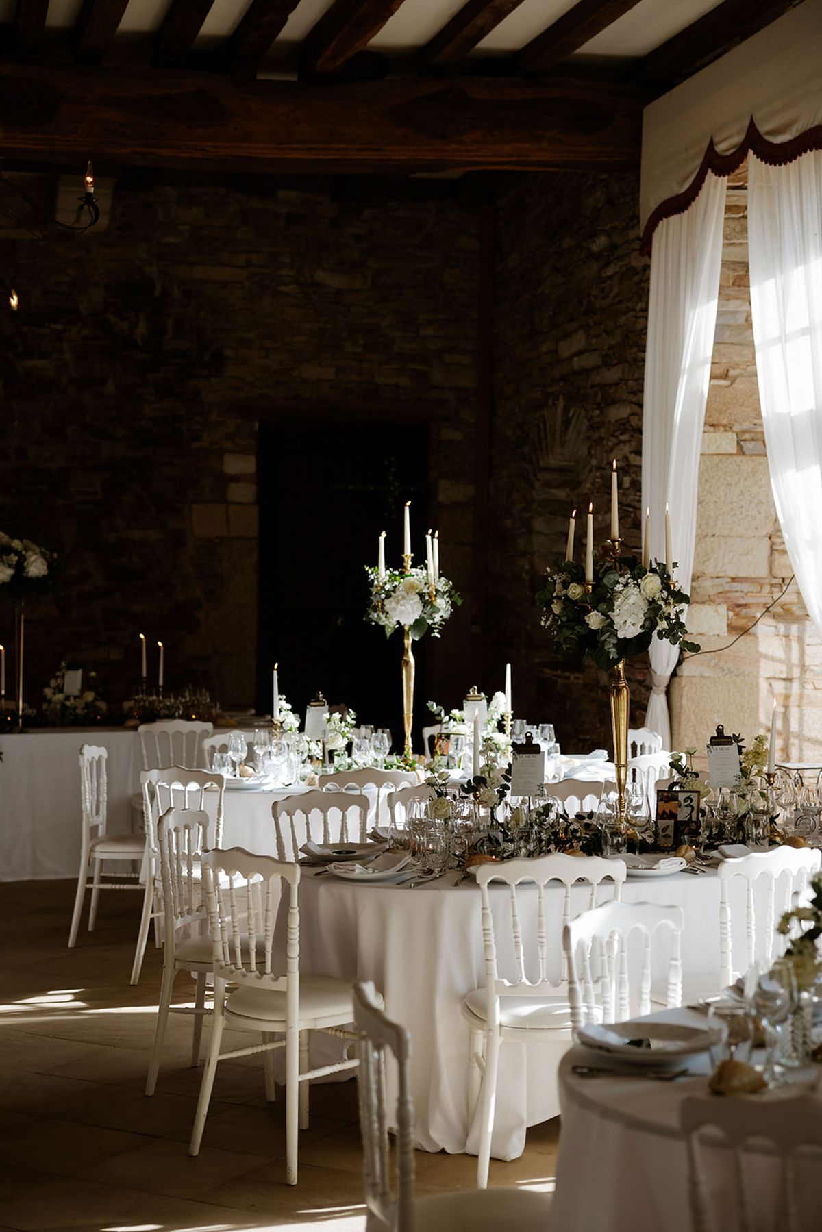 Indoor wedding reception setup photographed in a wide shot, showing round tables dressed in white floor-length linen cloths inside a stone-walled room with dark exposed wooden ceiling beams. The tables are surrounded by white Napoleon chairs and set with glassware, grey linen napkins, and place settings. Tall gold candelabras hold lit ivory taper candles and are topped with white floral arrangements featuring white roses, white hydrangeas, and greenery including eucalyptus. A trailing greenery and white floral runner decorates the table surface. Sheer white curtains with a burgundy scalloped trim hang at the window, allowing natural light to flood the space. Additional candlelit tables are visible in the background, maintaining the same white and gold decor palette. The overall styling theme is classic and formal with a green and white floral palette against gold metallic accents.