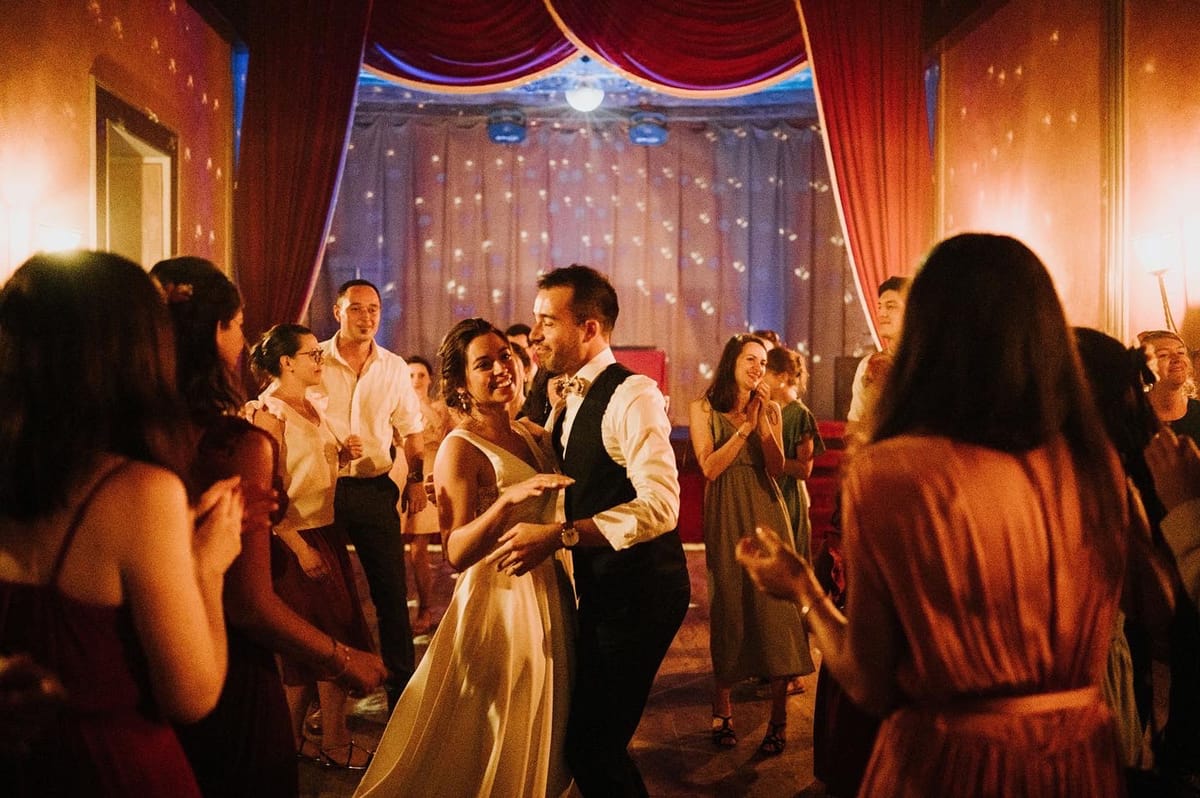 The bride and groom share a first dance at an indoor evening reception, surrounded by approximately 15–20 guests who are clapping and celebrating around them. The bride wears a simple ivory satin gown with a V-neckline, and the groom is dressed in a white shirt with a dark navy waistcoat and bow tie. The venue features deep red theatrical curtain draping along the back wall, warm amber wall lighting, and a sheer backdrop with scattered fairy lights or projected light spots, giving the room a theatrical, vintage ballroom atmosphere. The wide-angle shot is taken from within the crowd at floor level, placing the couple at the center of the frame with guests visible on all sides in the foreground and background.