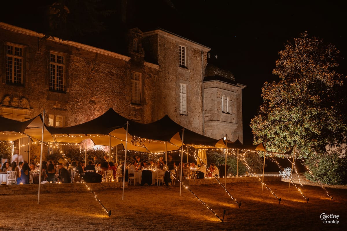 An outdoor evening wedding reception taking place on the grounds of a French château, photographed as a wide shot at night. Guests — approximately 60 to 80 people — are seated at round tables with white Chiavari chairs beneath a series of connected black stretch tents, with warm-toned fairy lights strung along the tent poles and across the ground leading toward the lawn. The tables are lit with candles, creating a warm amber glow across the dining area. The stone façade of the château is illuminated by uplighting, revealing multi-story architecture with arched windows and a rounded tower structure. The overall decor palette is warm gold and black, with the fairy light strands extending outward from the tent structure across the surrounding grounds. Potential venue feature image.
