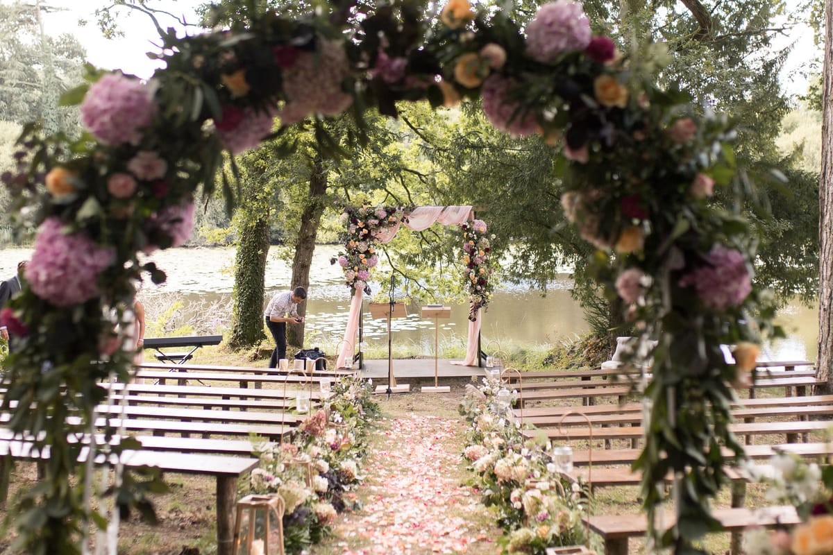 An outdoor ceremony setup photographed through a large circular floral arch in the foreground, which is densely decorated with mauve and lilac hydrangeas, peach and amber roses, deep burgundy blooms, and trailing greenery. The aisle runs through the center of the frame, lined with rose petals in blush and coral tones, low floral arrangements of mixed garden flowers, and gold lanterns placed at intervals along wooden pew-style benches on both sides. At the far end of the aisle, a rectangular altar arch draped with blush pink fabric panels is heavily decorated with matching floral arrangements in peach, mauve, burgundy, and ivory tones. A person — likely a vendor or musician — is visible near the altar adjusting equipment, with a keyboard instrument set up to the left. The ceremony is set on a flat grassy area beside a calm lake or pond, with mature trees framing the space. The overall decor palette is romantic garden-style with a mix of blush, mauve, peach, and burgundy florals against natural wood furniture. Wide establishing shot taken from behind the entrance arch.