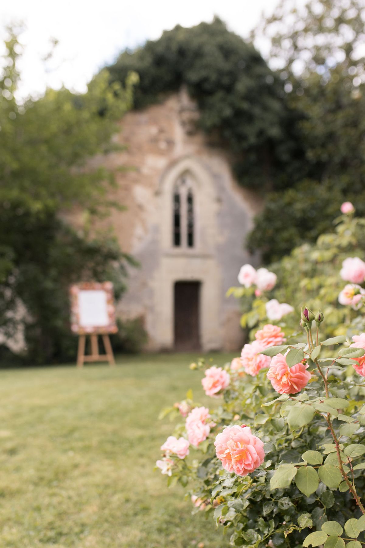 An outdoor detail shot taken in a garden setting, with a rose bush bearing fully open salmon-pink and blush roses in sharp focus in the foreground. In the soft-focus background, a rustic stone chapel or folly with a Gothic arched window and dark wooden doorway is visible across a lawn. A wooden easel holding a white framed wedding welcome sign with a floral-decorated border is positioned in front of the chapel entrance. The composition uses a shallow depth of field to draw attention to the roses while placing the venue and signage as contextual background elements. Potential venue feature image.