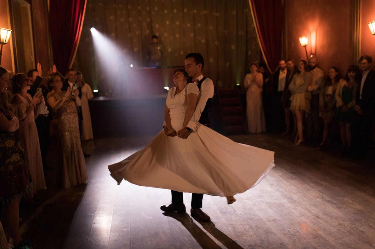 The couple's first dance is captured mid-spin in an indoor ballroom setting with warm amber wall sconces, deep red velvet curtains, and a gold-patterned backdrop. The bride wears a short-sleeved, knee-length white dress with a full, flared skirt that is caught mid-swirl, while the groom wears dark trousers, a white shirt, dark waistcoat, and a bow tie. A single spotlight from above illuminates the couple at the center of a polished parquet dance floor, with the DJ booth visible in the background. Approximately 20–25 guests line both sides of the dance floor, several applauding and recording on phones, dressed in formal evening attire in neutral and muted tones. The overall decor style is classic and theatrical, with rich warm lighting creating strong contrast against the darkened room. Wide shot taken from floor level.