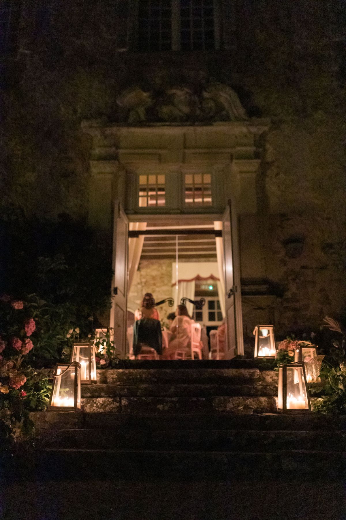 A nighttime outdoor shot looking up a stone staircase toward the open double doors of a French château, where a warmly lit reception is visible inside with several guests seated at tables. Tall glass-and-metal lanterns with lit candles are placed on each step flanking the entrance, providing the primary light source along the staircase. Floral arrangements in pink and coral tones are tucked among the lanterns on either side of the steps. Two figures in colorful dresses — one in teal, one in lighter pink — stand at the threshold facing inward toward the reception. The château facade features ornate carved stone detailing above the doorway, including decorative sculpted figures. The image is taken from ground level as a wide shot, with the dark exterior contrasting sharply against the amber glow from inside. Potential venue feature image.