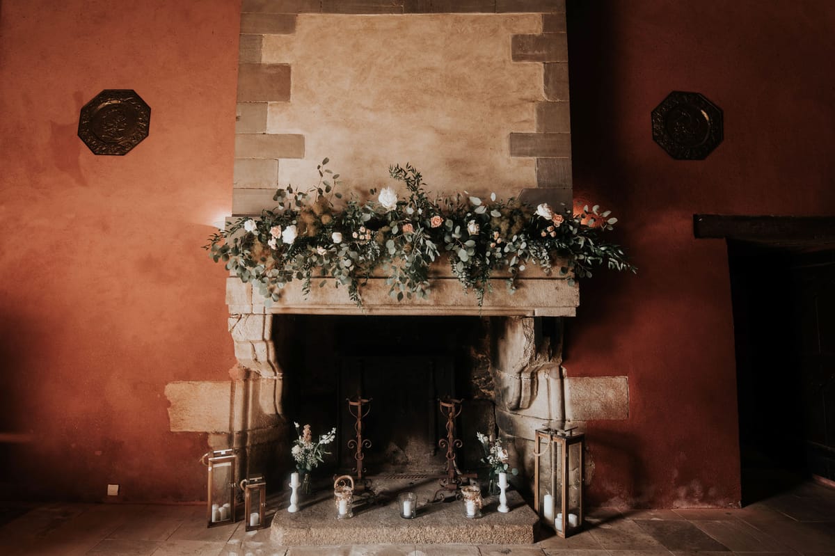 Detail shot of a large stone fireplace decorated for a wedding, set inside a room with terracotta-red painted walls and two octagonal decorative metal plaques mounted symmetrically on either side. The fireplace mantel is dressed with a lush floral garland featuring eucalyptus, white roses, peach roses, and what appears to be dried pampas-style grass, trailing loosely over the edges. Inside the fireplace hearth, the styling includes two tall white pillar candles on wrought iron candlestick holders, several glass hurricane lanterns with white candles, small bud vases with white flowers, and gold-toned lanterns flanking the exterior. The overall decor palette is warm — terracotta, peach, white, and sage green — with a rustic yet polished styling approach. Potential venue feature image.
