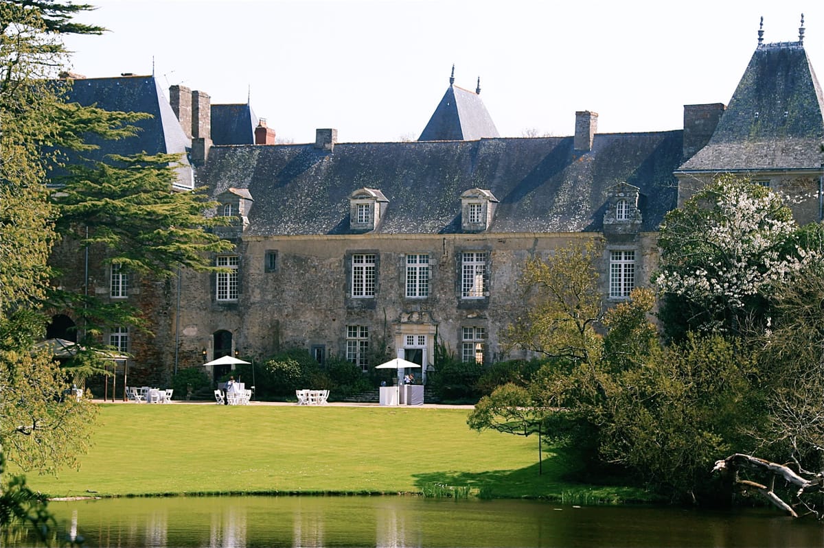 Wide exterior shot of a French château wedding venue captured from across a ornamental pond, showing the full facade of a large stone manor house with slate mansard roofs, dormer windows, and pointed corner towers. Along the terrace in front of the building, white chairs and tables are arranged under two open white market umbrellas, with what appear to be a small number of people — likely event staff or early-arriving guests — setting up or attending a cocktail-style outdoor area. A white draped bar or display table is visible near the central entrance doors. The setup suggests an outdoor cocktail hour or reception area styled with a clean white decor palette against the historic stone architecture. Potential venue feature image.