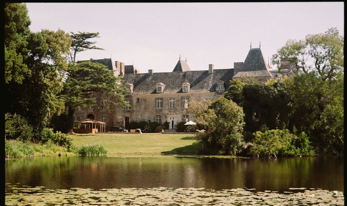 Wide exterior shot of a French château venue photographed from across a pond covered with lily pads. The multi-story stone building features a slate mansard roof with pointed towers, dormer windows, and multiple chimneys, consistent with classic French manor architecture. On the grounds in front of the château, a wooden open-sided pavilion structure and at least one white market umbrella are visible, suggesting event setup in progress, along with a dark-colored vehicle parked nearby. The image was shot on film, giving it a warm, slightly faded tone with characteristic film grain and dark vignetting along the edges. Potential venue feature image.