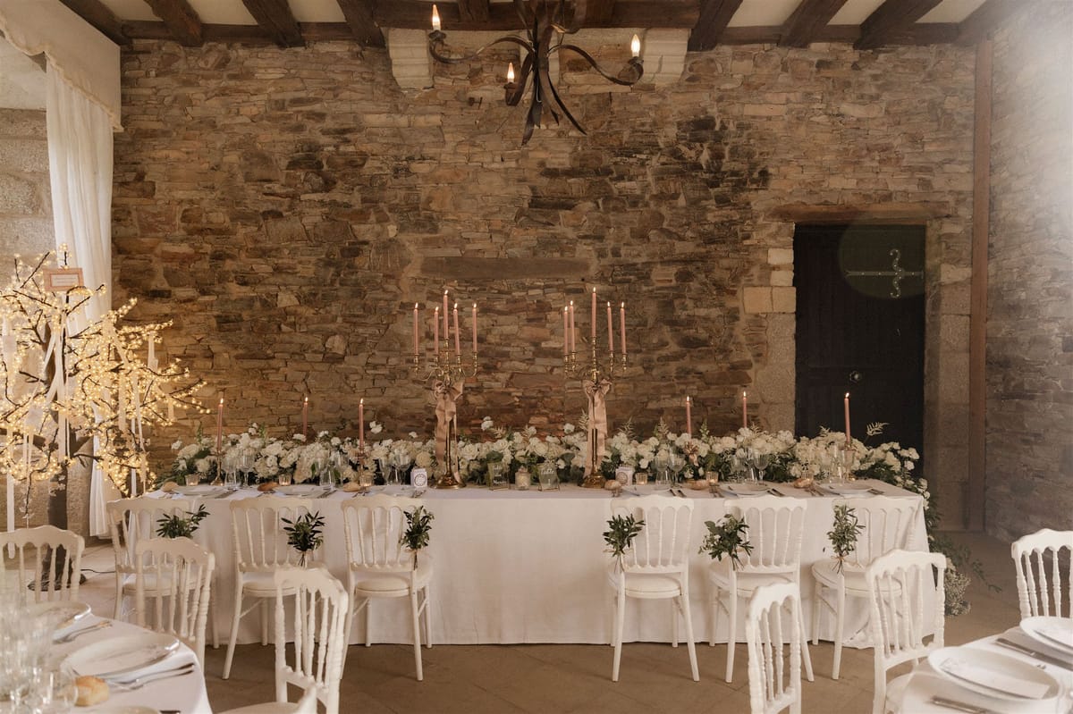 A wide shot of a decorated wedding reception head table set inside a rustic indoor venue with exposed stone walls and wooden ceiling beams. The long rectangular table is dressed in a white linen cloth and lined with white Napoléon chairs, each adorned with small greenery bundles tied with ribbon. The table centerpiece features a continuous low floral runner of white hydrangeas and mixed greenery, flanked by two tall gold candelabras holding blush-pink taper candles, with additional single blush taper candles spaced along the runner. Place settings include white plates, glassware, and gold accents. To the left, two illuminated decorative branch trees wrapped in warm white fairy lights add ambient light. An ornate wrought-iron chandelier hangs from the ceiling above. The overall decor palette is white, gold, and blush pink, with a classic French rustic styling. Potential venue feature image.