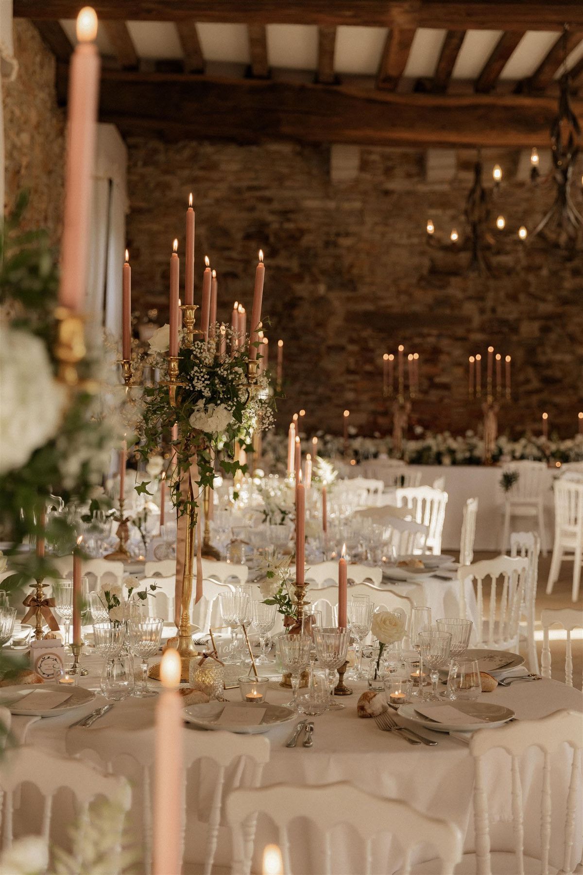 Indoor wedding reception tablescape detail shot taken in a rustic barn or stone-walled venue with exposed wooden beam ceiling and warm Edison bulb string lighting overhead. The round tables are dressed in white linen and set with crystal glassware, grey charger plates, silver cutlery, and small place cards with ribbon details. Tall brass candelabras serve as centerpieces, holding lit dusty rose taper candles and decorated with trailing ivy, white hydrangeas, and white gypsophila. Additional individual brass candlestick holders with matching dusty rose tapers are placed around each table. The overall decor palette is white, gold, and dusty rose with green foliage accents, styled in a classic rustic-romantic aesthetic. White Napoleon-style chairs surround the tables, and multiple similarly decorated tables are visible extending into the background of the room.