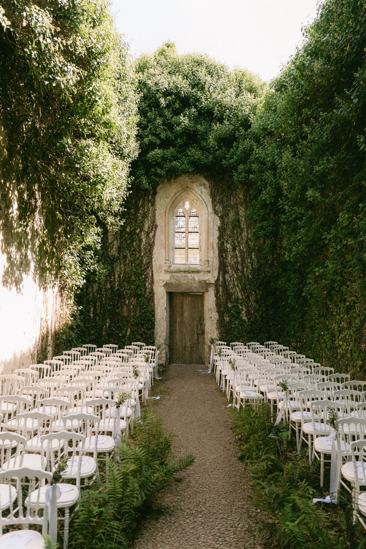 An outdoor wedding ceremony setup photographed from the back of the aisle looking toward the altar, with no guests or couple present. Approximately 20 rows of white Napoleon-style chairs with white cushions are arranged in two sections on either side of a gravel aisle, with potted ferns lining the base of each row. The focal point is an ancient stone wall almost entirely covered in climbing vines and ivy, featuring a tall Gothic arched window above a weathered wooden door. The setting has a classic, garden-ruin aesthetic with the ivy-covered walls creating a natural enclosure around the ceremony space. Wide shot composition emphasizing the symmetry of the chair layout and the architectural stone backdrop. Potential venue feature image.