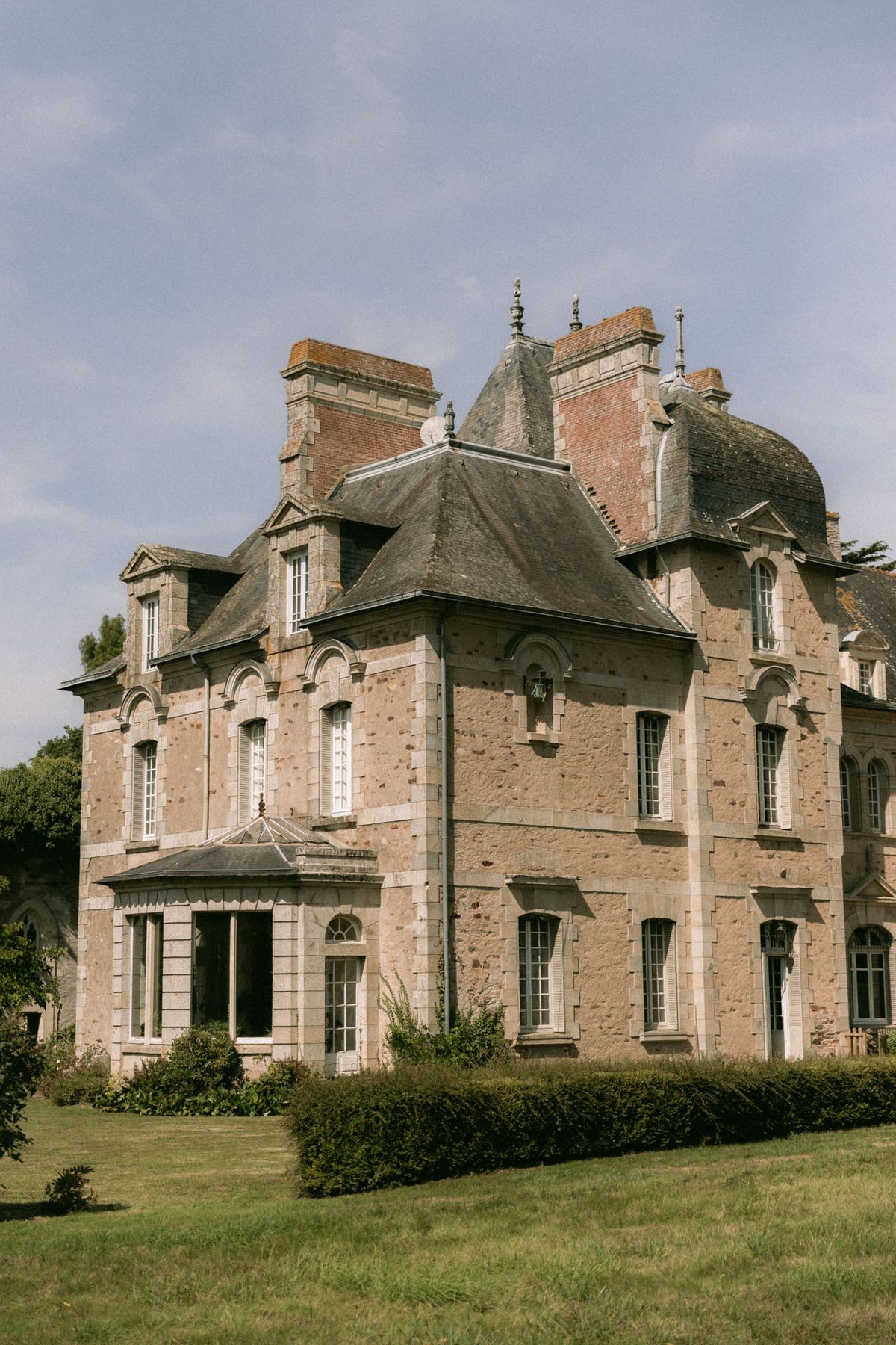 An outdoor wide shot of a French château photographed from the grounds, with no people visible. The building is a multi-story structure built in warm pinkish-beige granite stone with cream-colored window surrounds, white shutters, and arched window detailing. Architectural features include steeply pitched dark slate roofs, conical turret tops with decorative iron finials, multiple brick chimneys, and a glass-roofed conservatory extension on the lower left facade. The foreground includes a trimmed hedge border and a well-maintained lawn. Potential venue feature image.