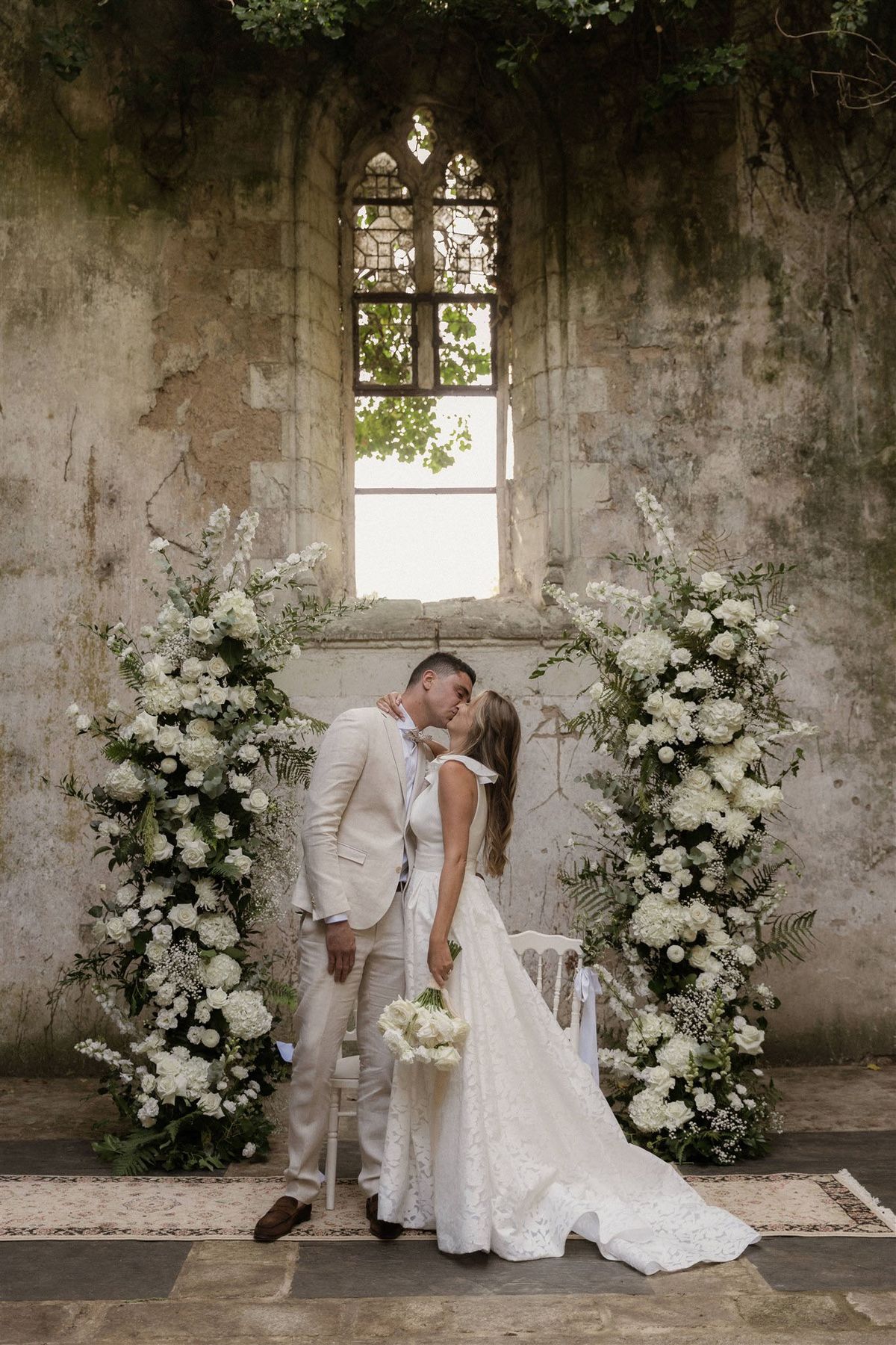 A bride and groom share their first kiss during or immediately after a ceremony, standing before two tall floral columns framing a Gothic arched window in a roofless stone ruin. The bride wears a white lace gown with a train and holds a white tulip bouquet with trailing white ribbon, while the groom wears a cream linen suit with brown loafers and a pale bow tie. The floral installations are composed of white hydrangeas, white roses, baby's breath, white delphiniums, and green ferns arranged in full, floor-to-ceiling columns. A vintage-style patterned rug is laid on the stone floor beneath them, and a white wooden chair is visible behind the couple. The setting is an open-air ruined chapel or historic stone structure with heavily weathered walls and climbing greenery overhead, styled with a romantic white-and-green palette. The shot is a full-length portrait taken from a slight distance, centering the couple symmetrically between the floral columns.