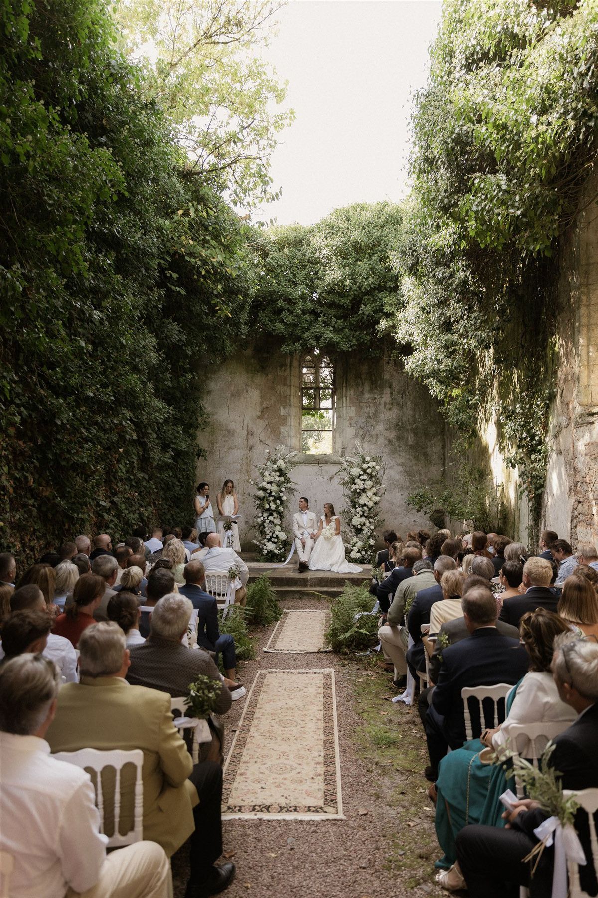 An outdoor wedding ceremony taking place within the roofless ruins of a stone chapel or church, with ivy and climbing greenery covering the tall stone walls on both sides. The couple — the groom in an ivory or off-white suit and the bride in a white gown holding a white bouquet — are seated on raised steps at the altar, facing a speaker or officiant who addresses the guests. The altar is framed by two large floral arrangements composed of white blooms and lush greenery forming an arch, set against the weathered stone wall with a gothic-arched window. Approximately 80–100 guests are seated on white chairs arranged in two sections on either side of an aisle lined with layered vintage-style rugs and low greenery, possibly fern fronds. The ceremony aesthetic is romantic and garden-inspired with an all-white and green floral palette. Wide shot taken from the back of the aisle looking toward the altar. Potential venue feature image.