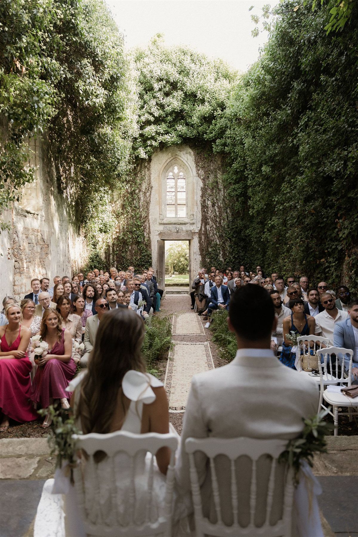 An outdoor wedding ceremony taking place within the roofless ruins of a stone chapel or abbey, its tall ivy-covered walls forming a natural corridor open to the sky. The bride and groom are seated at the foreground in white painted chairs decorated with greenery garlands, their backs to the camera, facing approximately 80–100 seated guests arranged in rows along both sides of a central aisle runner bordered by fern fronds. The bride wears a white dress with a large bow detail at the back, and the groom wears a white suit jacket. A gothic arched window opening remains intact in the far stone wall, framed by dense climbing vines, with a doorway below it providing a view into a garden beyond. Guest attire includes a raspberry-pink bridesmaid dress, a fuchsia dress, and a mix of smart-casual summer outfits. The ceremony styling is minimal and natural, with white chairs and greenery accents against the historic ruined architecture. Wide shot taken from slightly elevated behind the couple, capturing the full length of the ceremony space. Potential venue feature image.